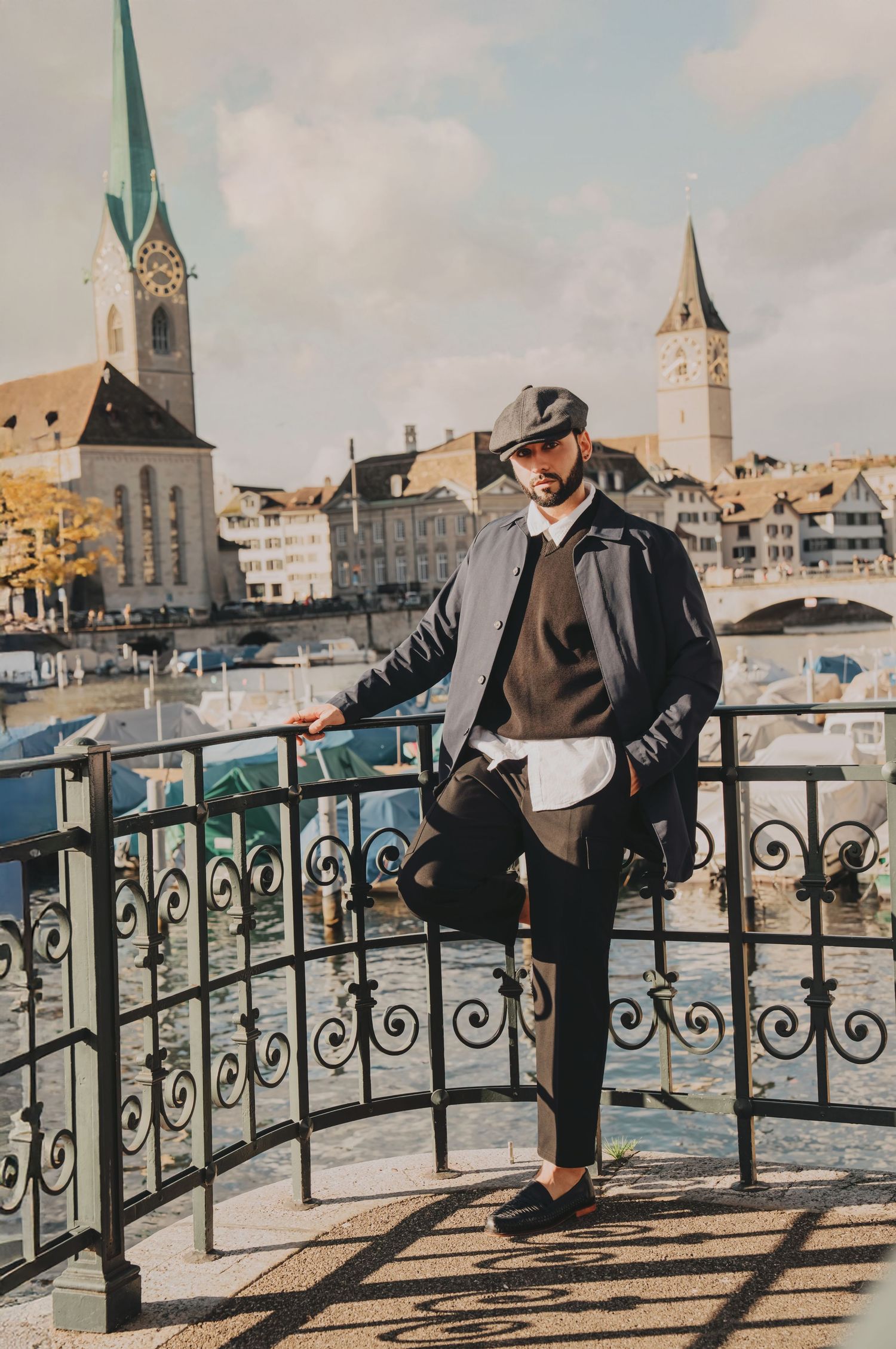 Stylish man in flat cap and layered outfit poses on ornate iron railing by Zurich river with church towers backdrop.