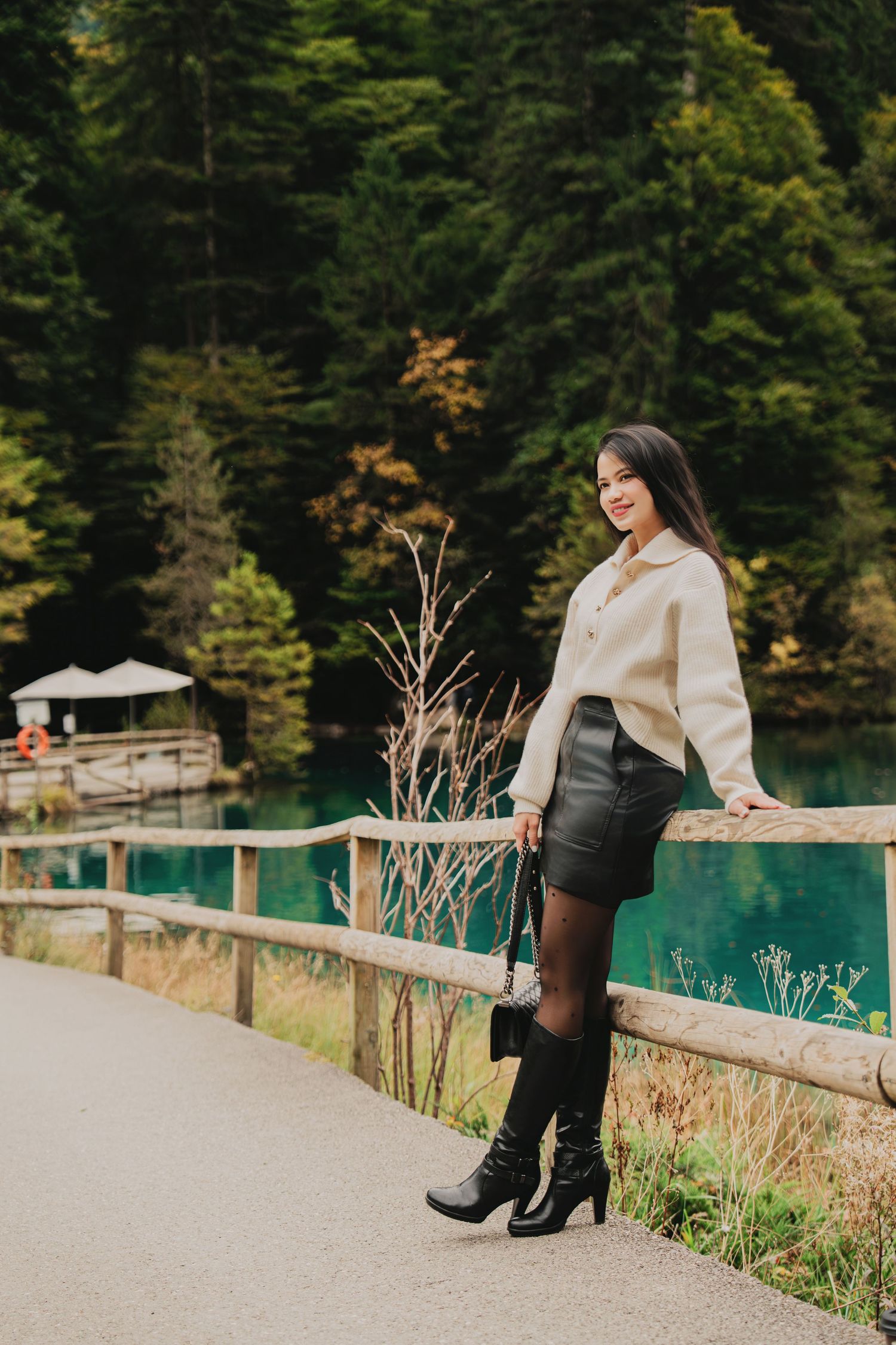 Fashionable woman in white sweater and black leather skirt poses by turquoise forest lake.