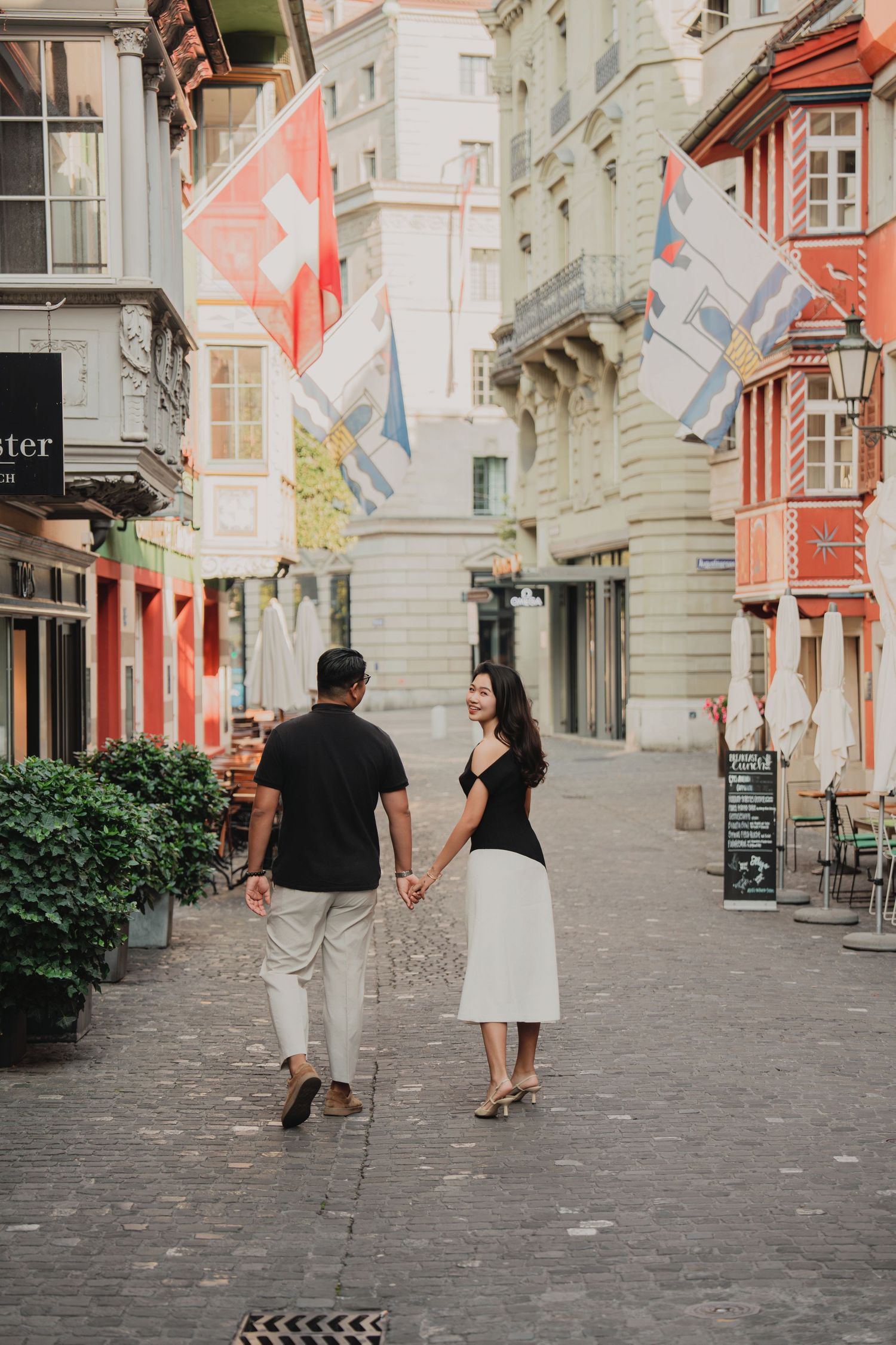 Couple holding hands walking through a vibrant Swiss old town street lined with colorful buildings and flags.