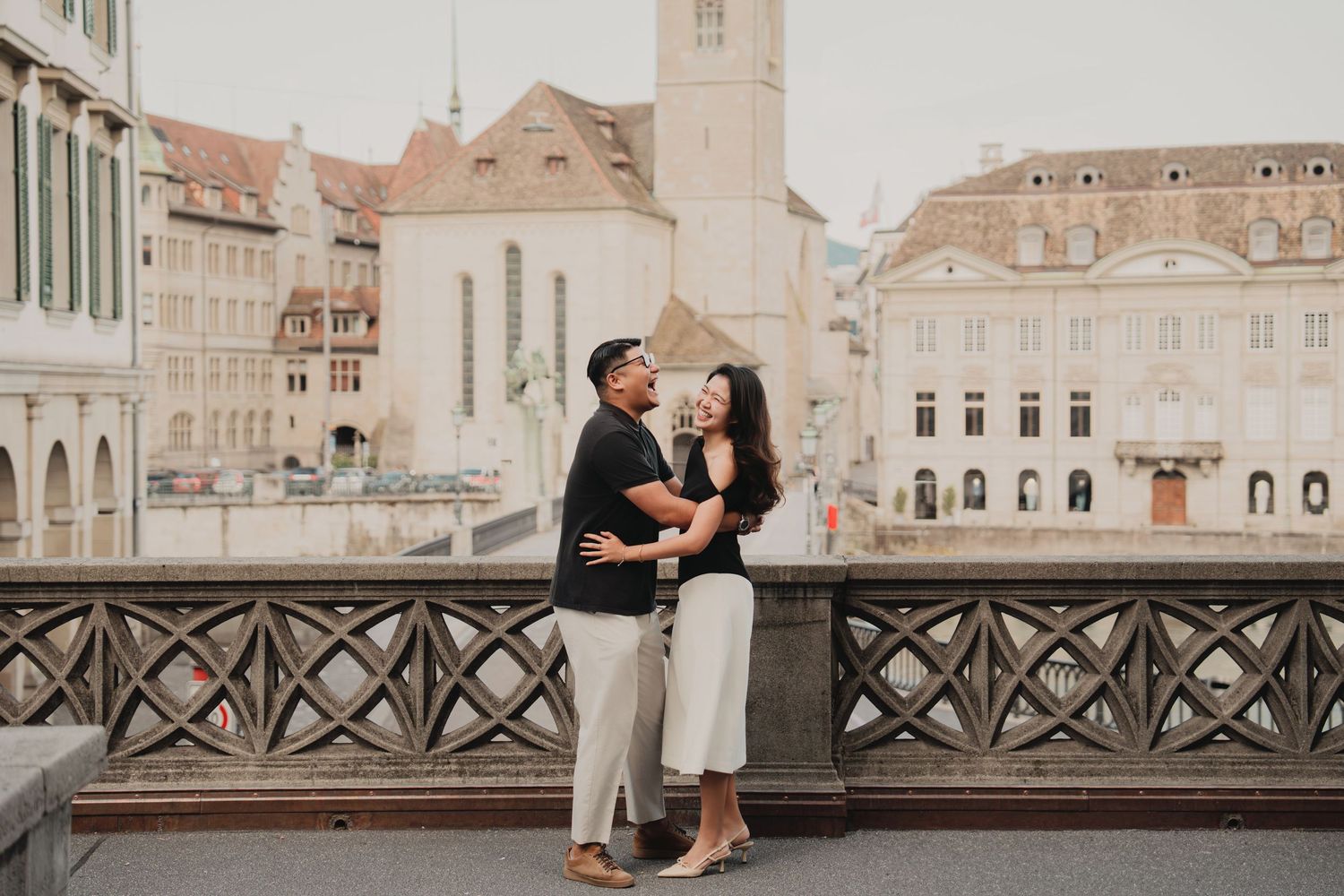 Couple hugging and smiling on a Zurich bridge with historic European buildings in the background.