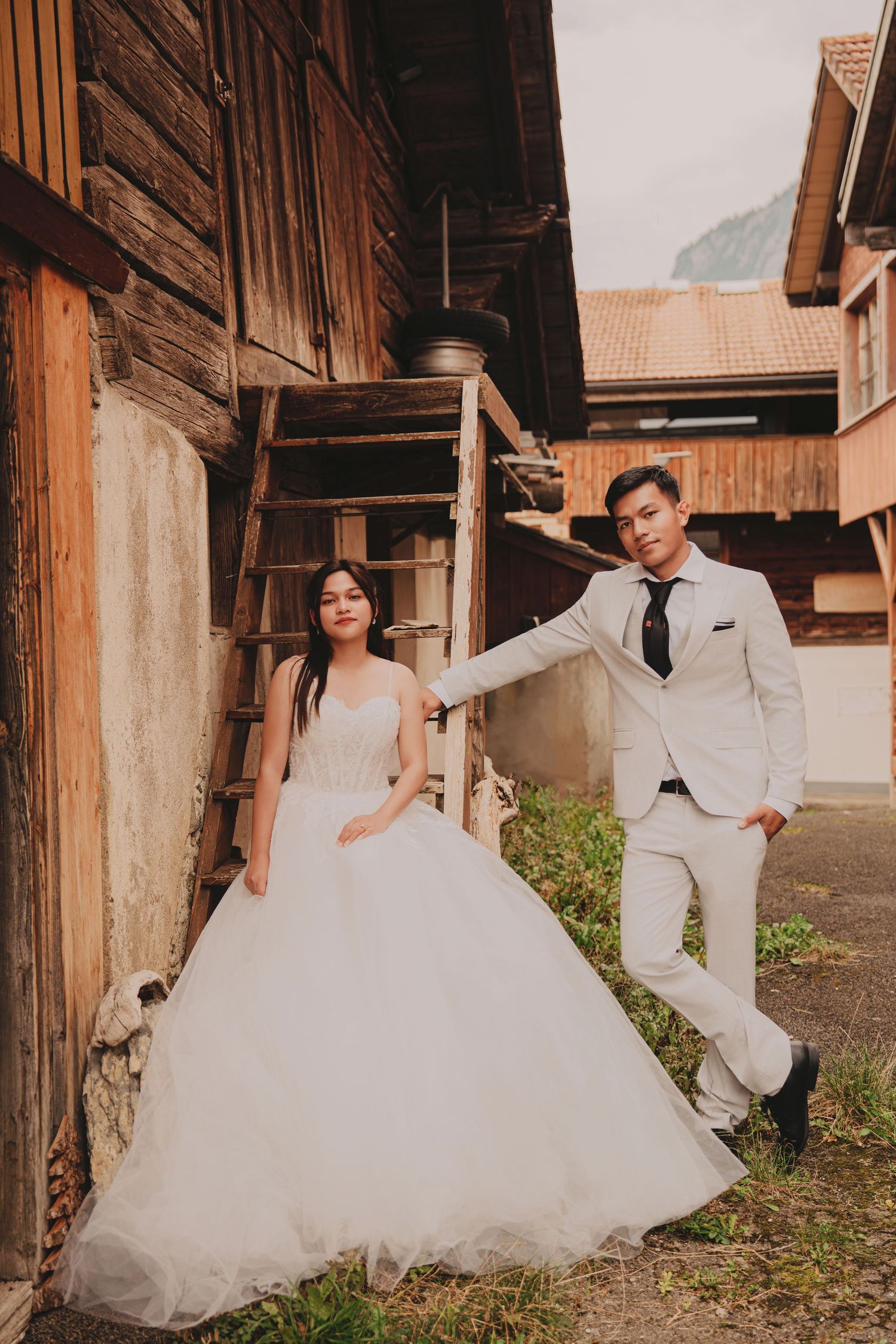 Elegant bride and groom strike a pose beside weathered wooden barn stairs in wedding attire.