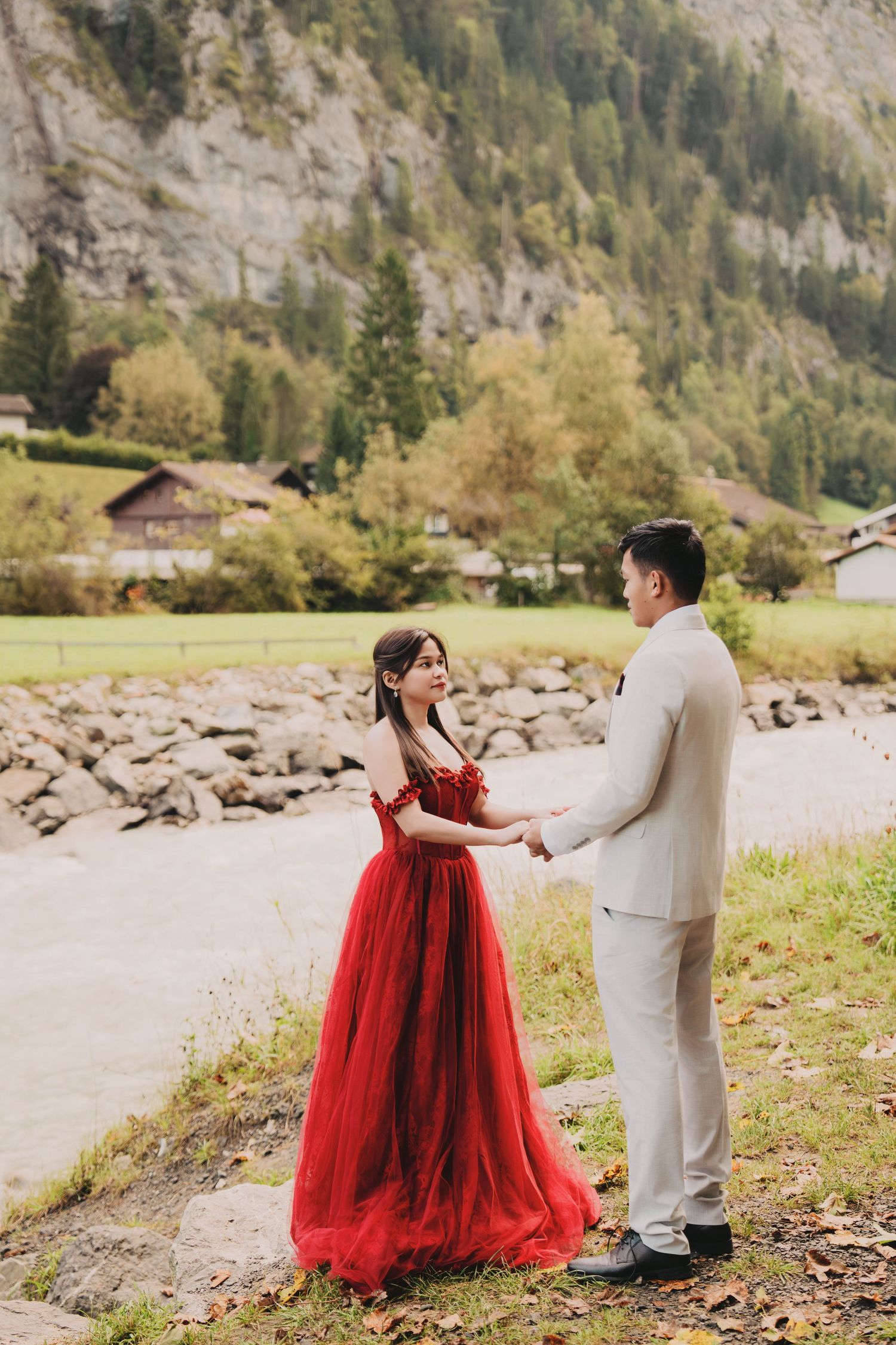 Asian couple holding hands in scenic Swiss alpine setting, bride wearing elegant red tulle gown.