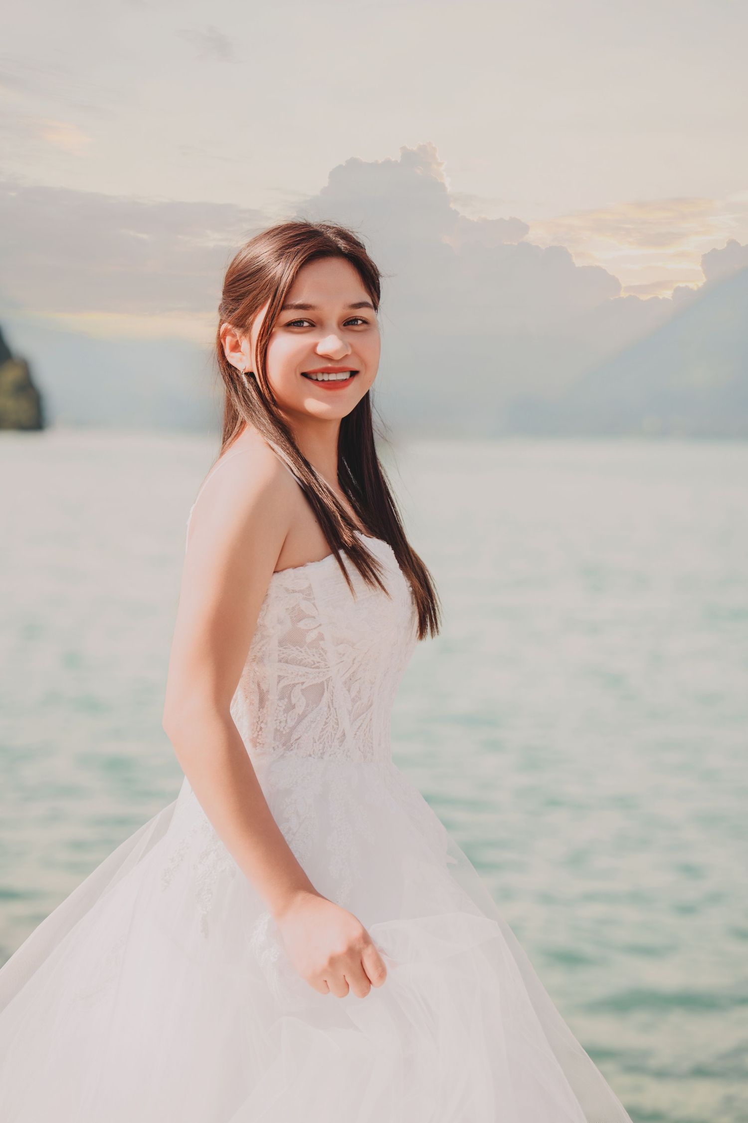 Joyful bride in white lace wedding dress smiles by a calm turquoise lake with dramatic cloudy mountain sky.