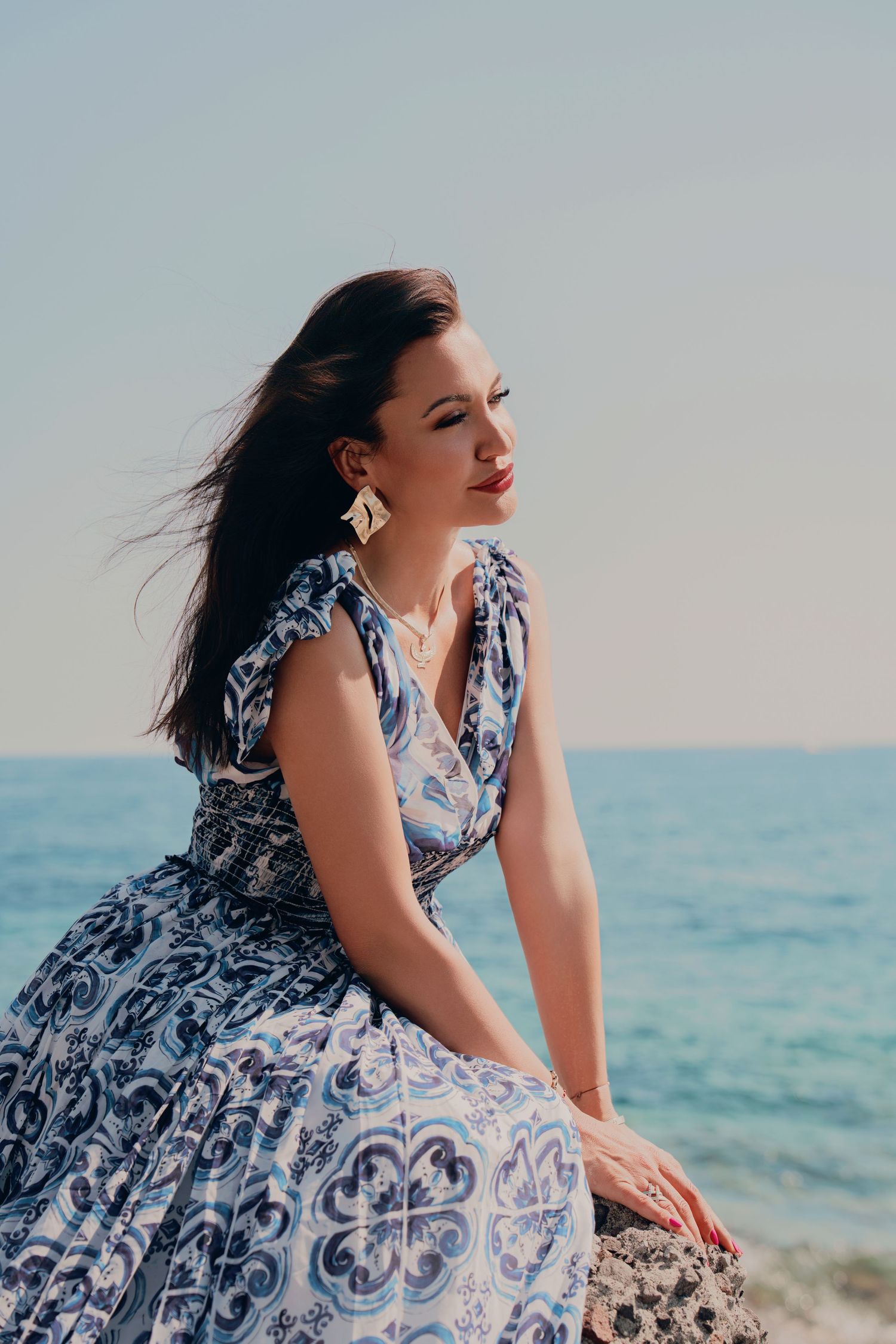 Stylish woman in blue Majolica print dress with gold earrings posing on rocks by the ocean.