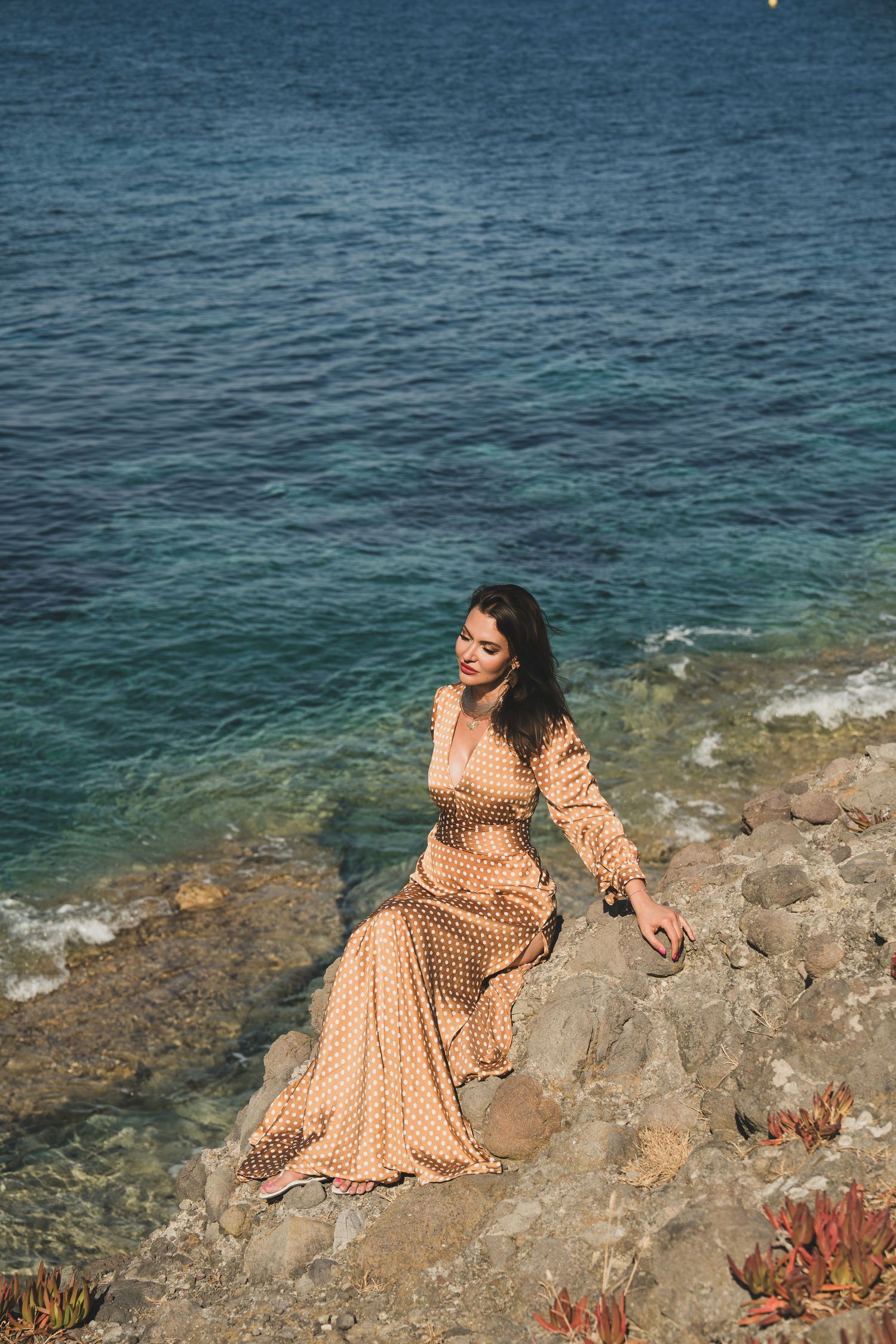 Smiling brunette in gold polka dot dress seated on rocky cliff above blue ocean.