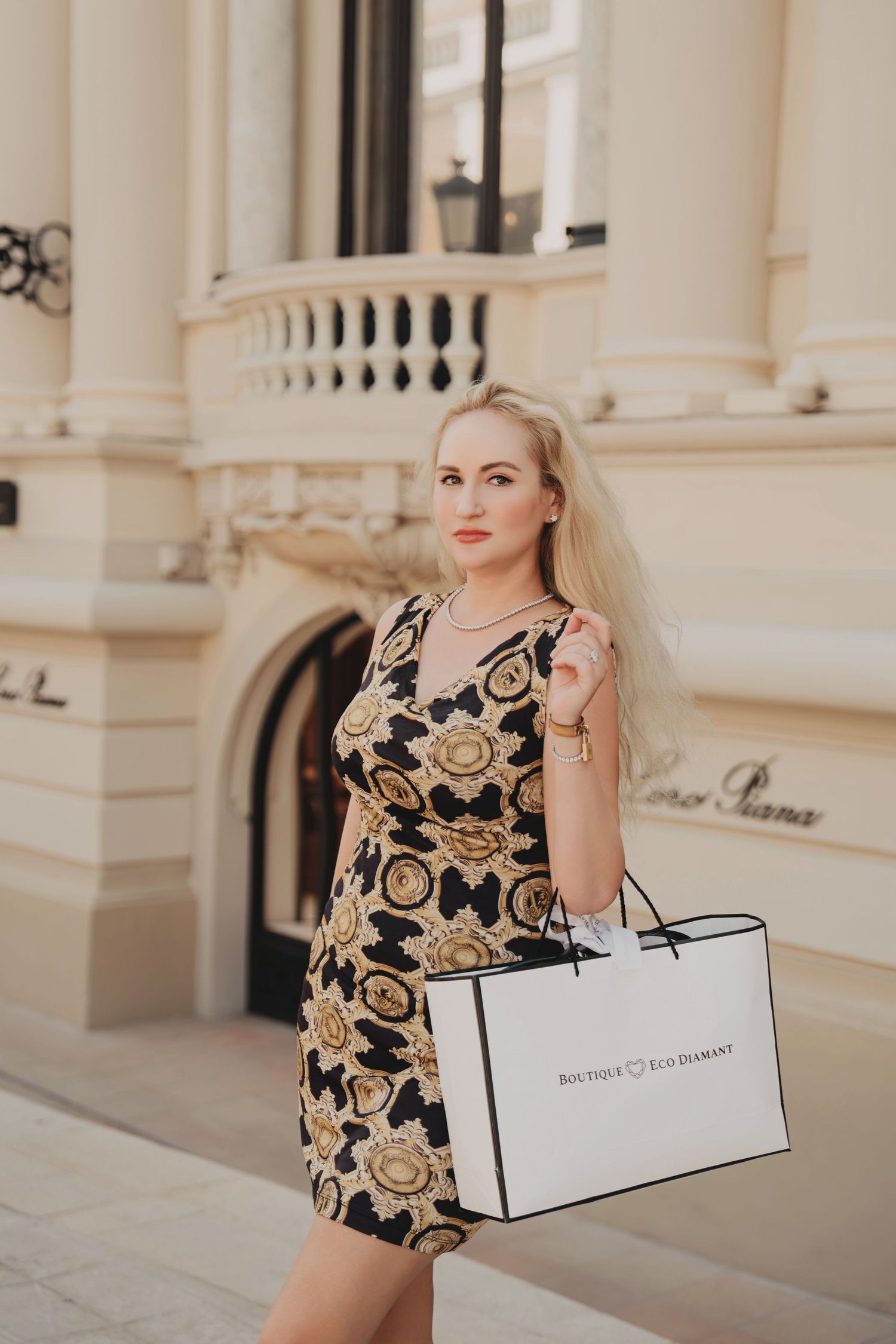 Fashionable blonde holding Boutique Eco Diamant shopping bag wearing gold print mini dress.