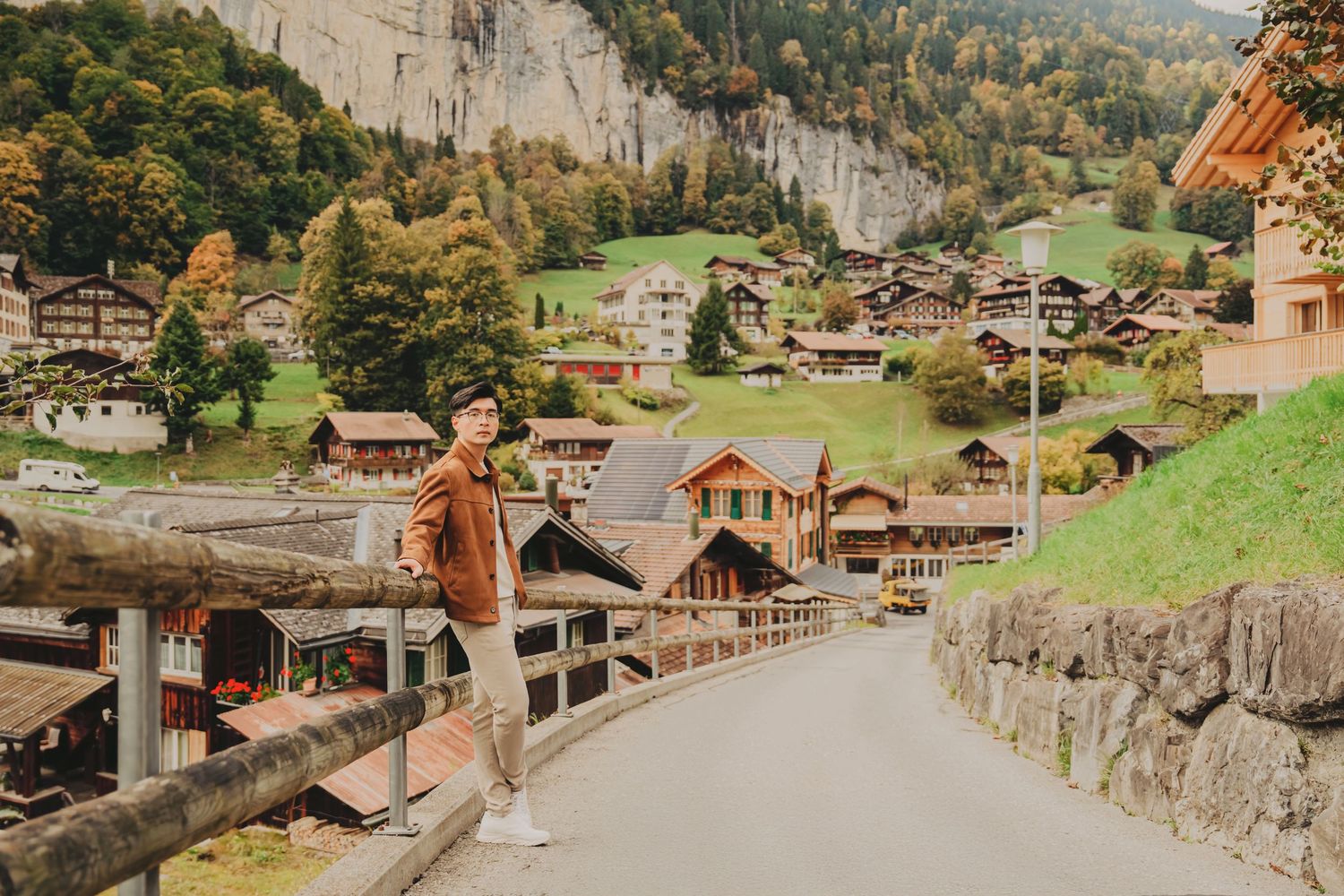 Young man in brown jacket posing on a bridge with a scenic Swiss alpine village and mountain backdrop.