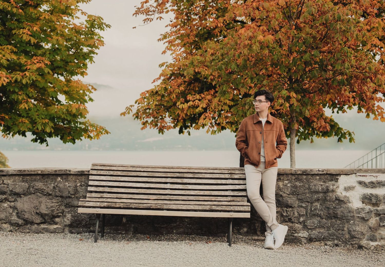Young man in a brown jacket leaning against a wooden bench framed by colorful autumn trees near a lake.