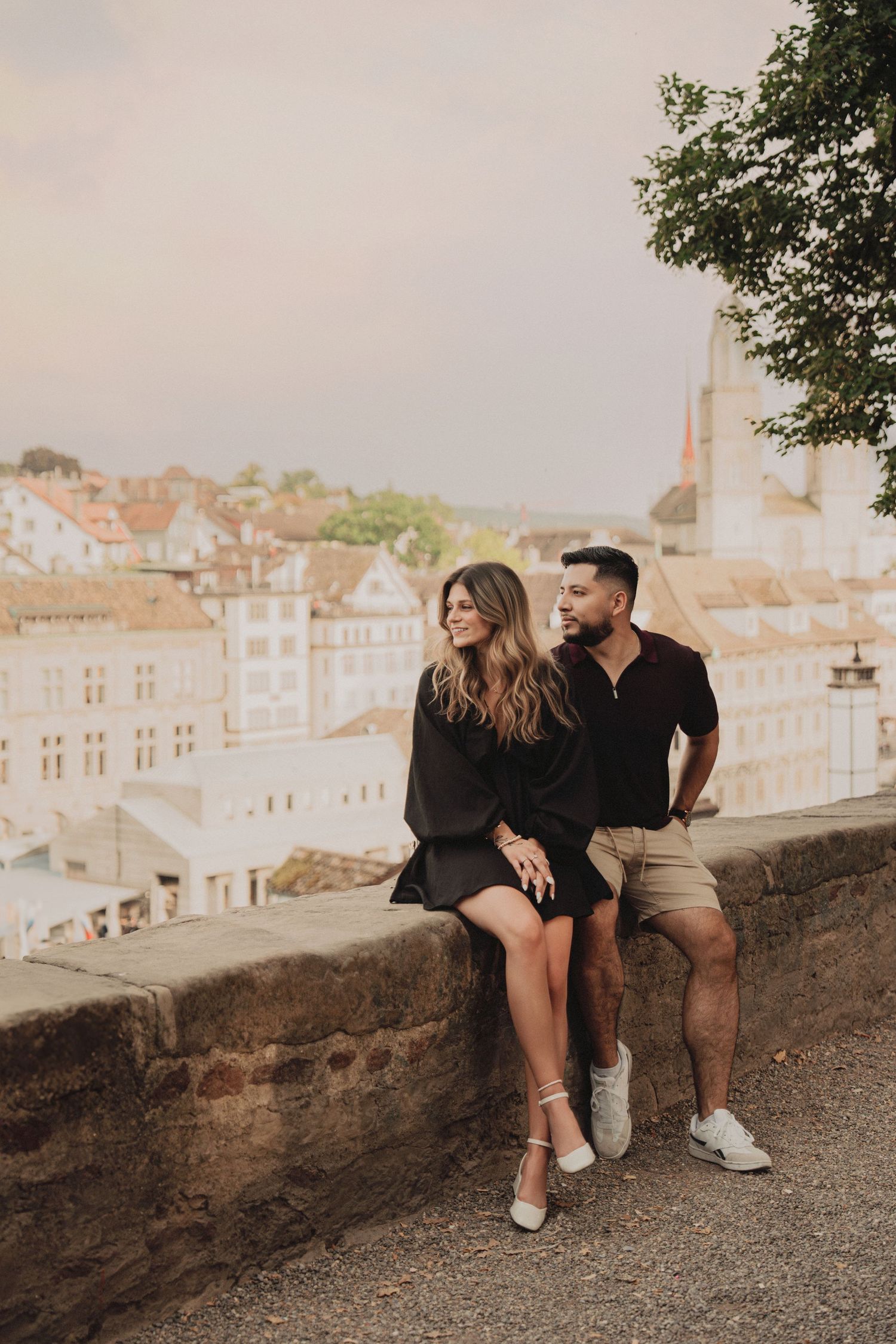 Couple relaxing on a historic stone wall overlooking the charming red-roofed buildings of Zurich's old town.