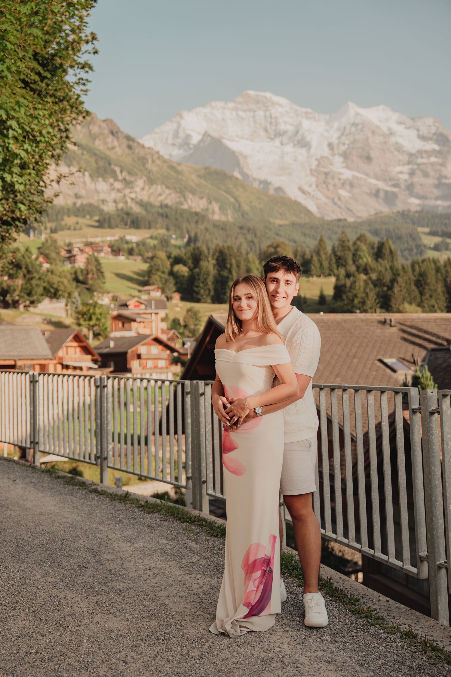 Young couple posing together on a mountain road with stunning Swiss Alps and village backdrop in golden hour light.
