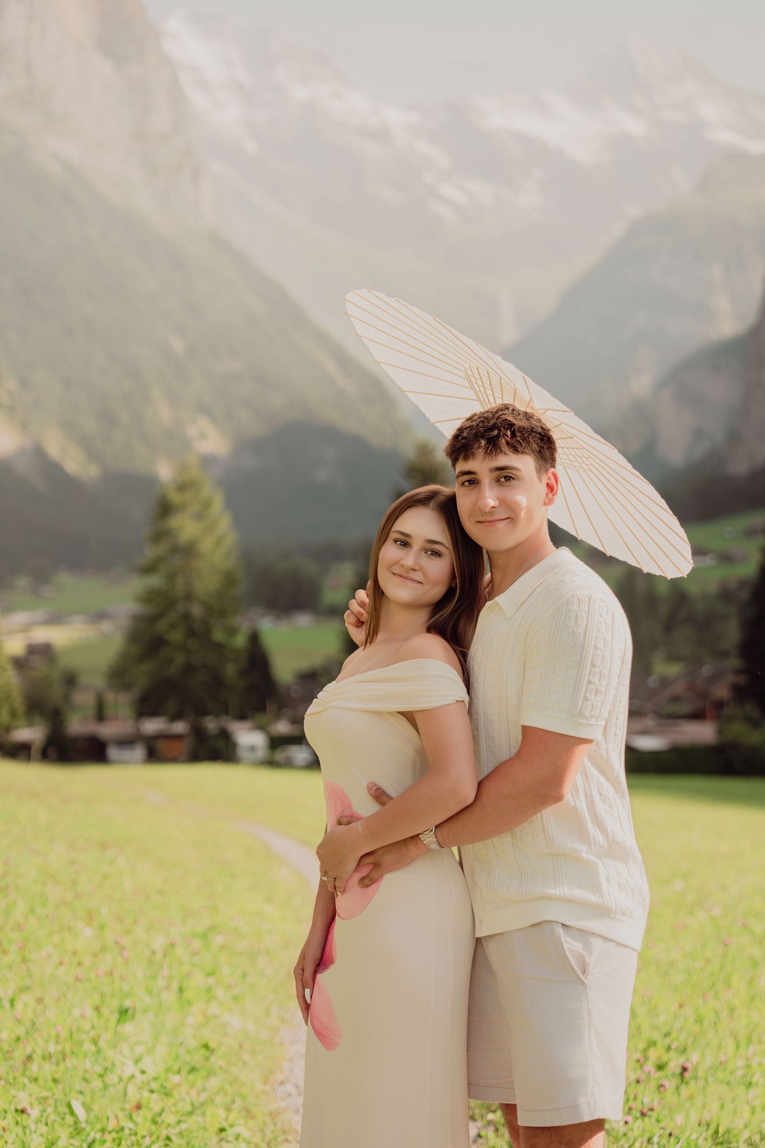Loving couple embracing on green meadow with white parasol and breathtaking Swiss mountain valley in the background.