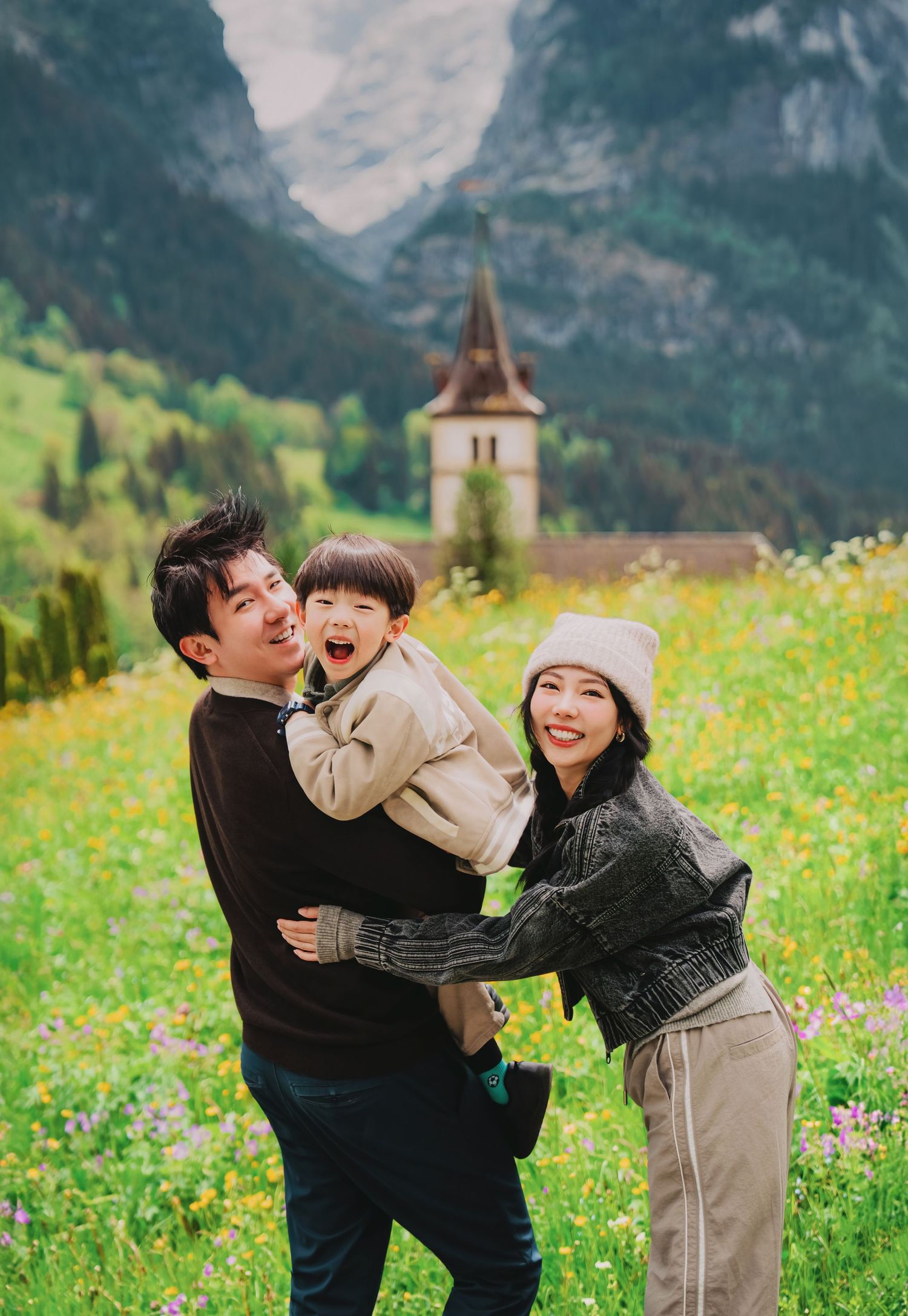 A happy Asian family poses with their laughing toddler in a Swiss Alpine meadow with a church steeple behind.