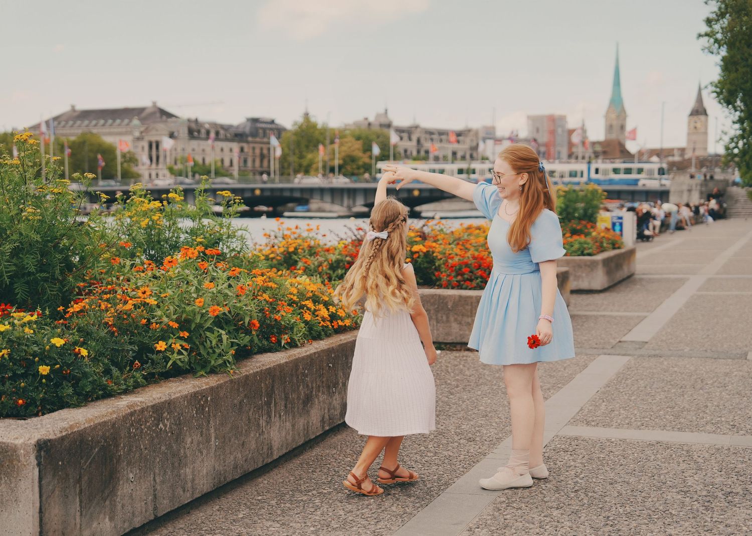 Two young girls in dresses admiring colorful flowers along Zurich riverside promenade.