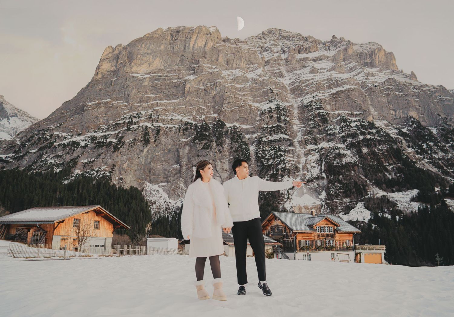Couple standing in snowy field with crescent moon above rugged Dolomites mountain and chalet village.