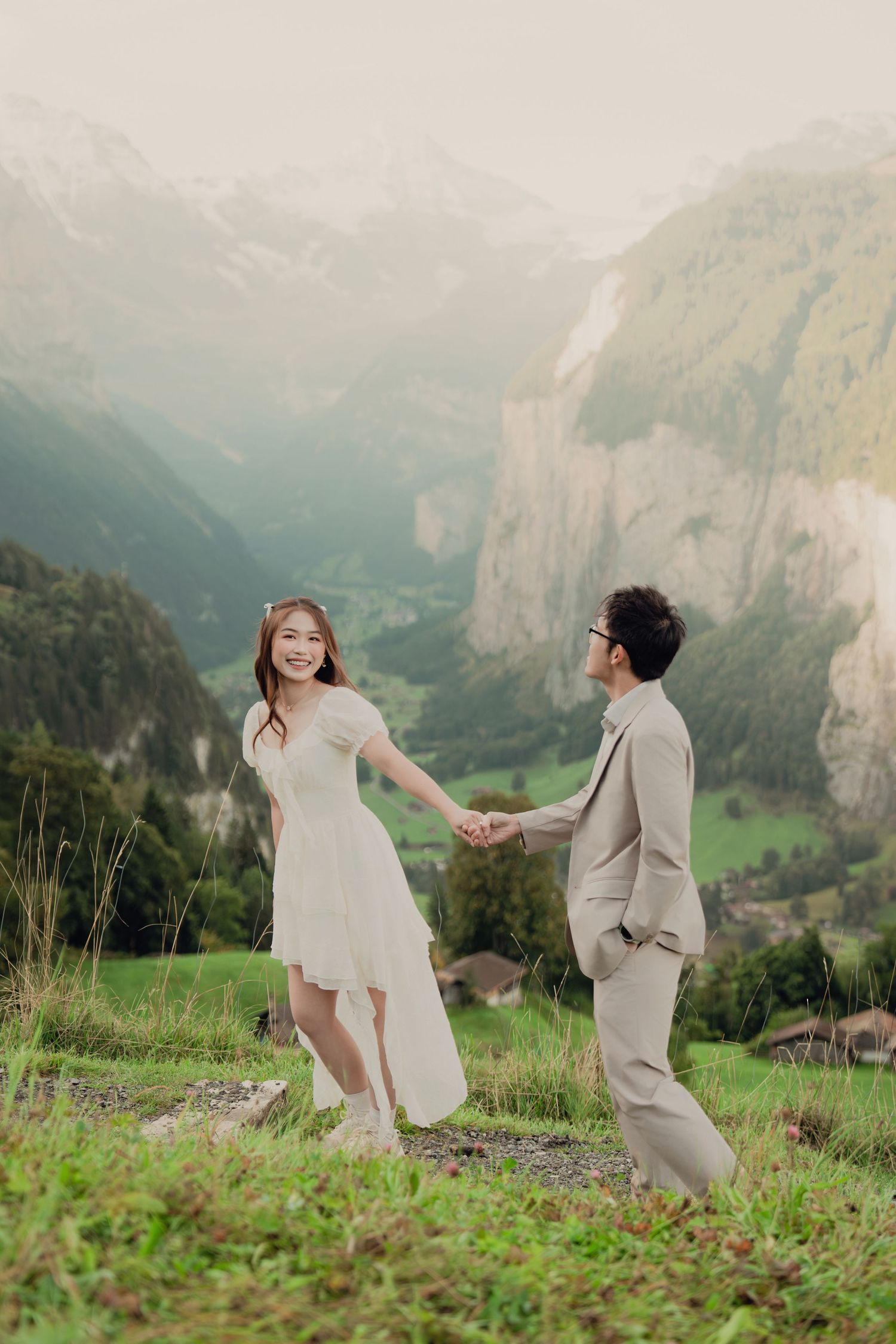 Joyful couple holding hands on green hillside with stunning Lauterbrunnen valley and mountains behind them.