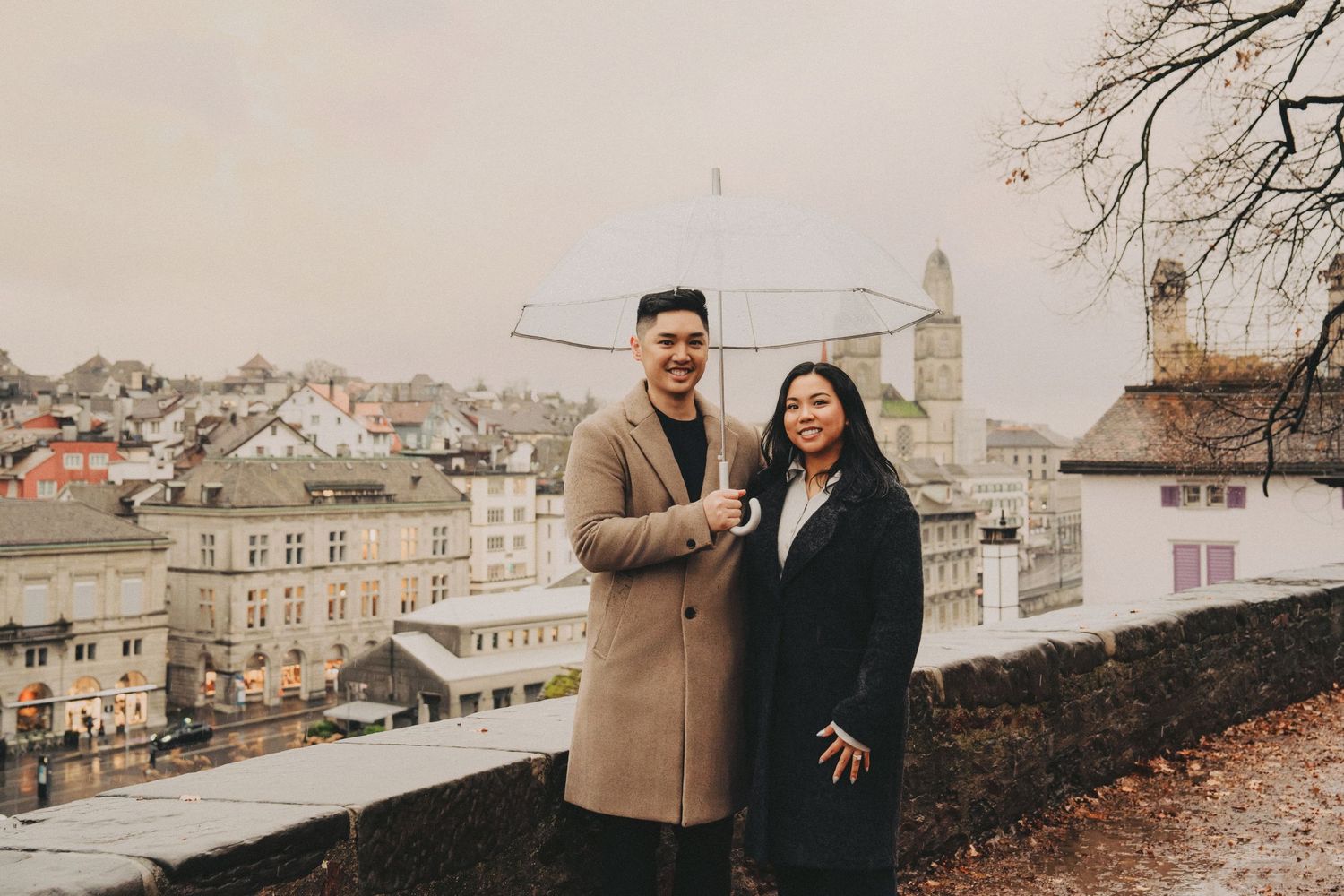 Smiling couple posing with a clear umbrella on a rainy day with Zurich old town behind them.