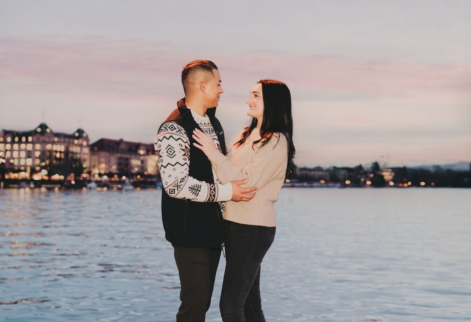Couple gazing at each other near a waterfront with softly lit buildings at dusk.