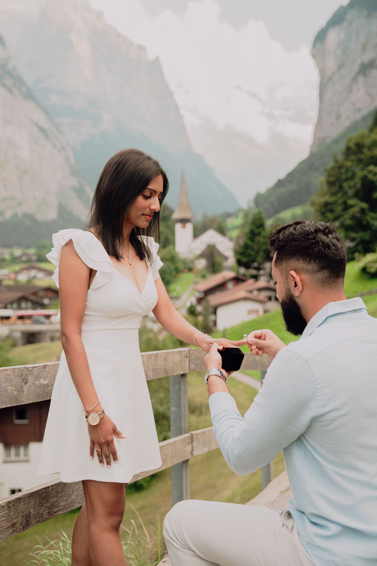 Man on one knee presents engagement ring box to smiling woman in white dress in Swiss Alps.