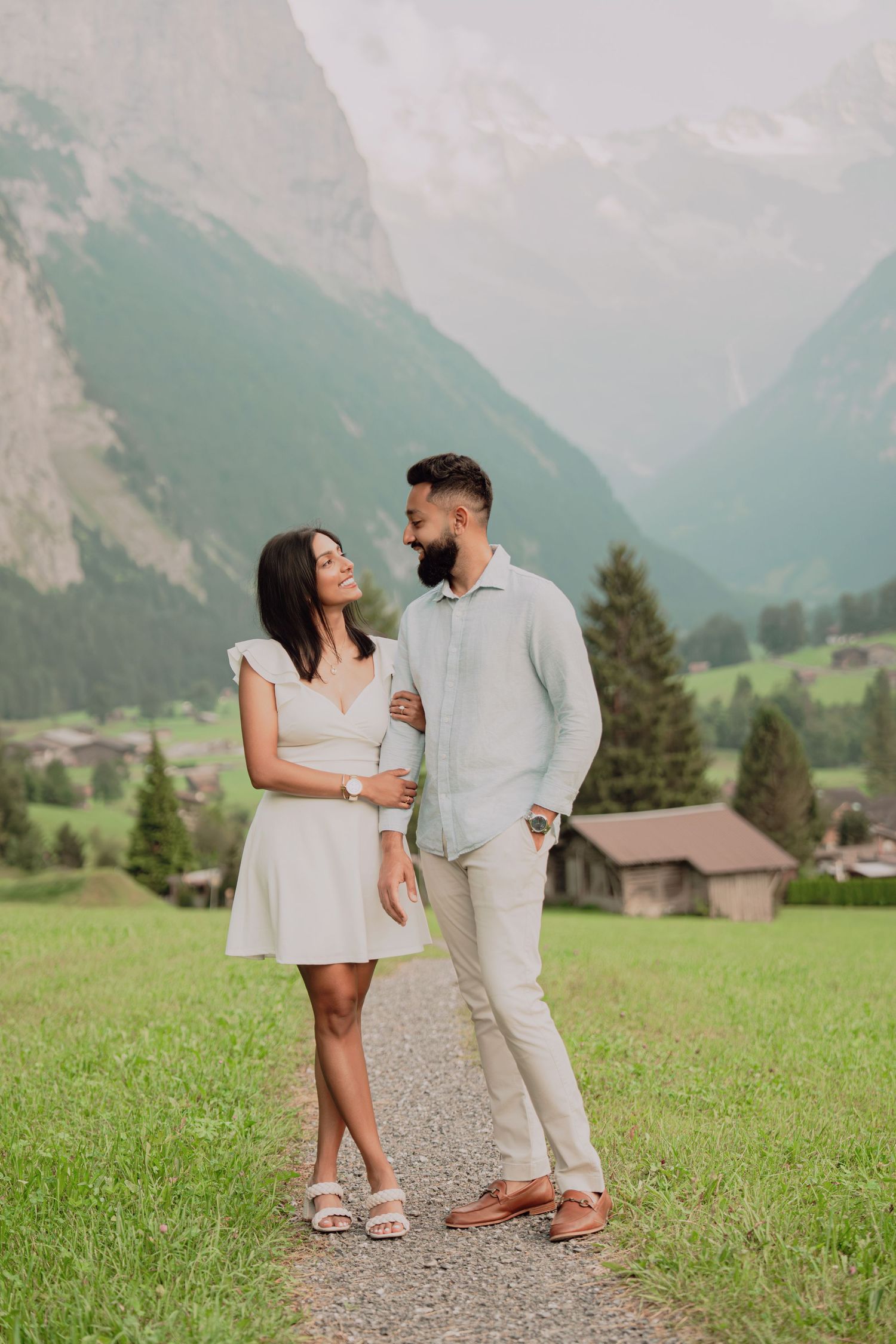 Romantic couple embracing on gravel path with lush Swiss alpine valley, green meadows and mountain peaks behind them.