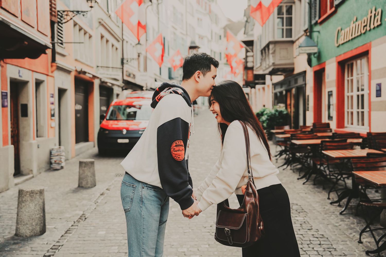 A romantic couple leans in for a kiss while holding hands on a flag-lined cobblestone street in Zurich.