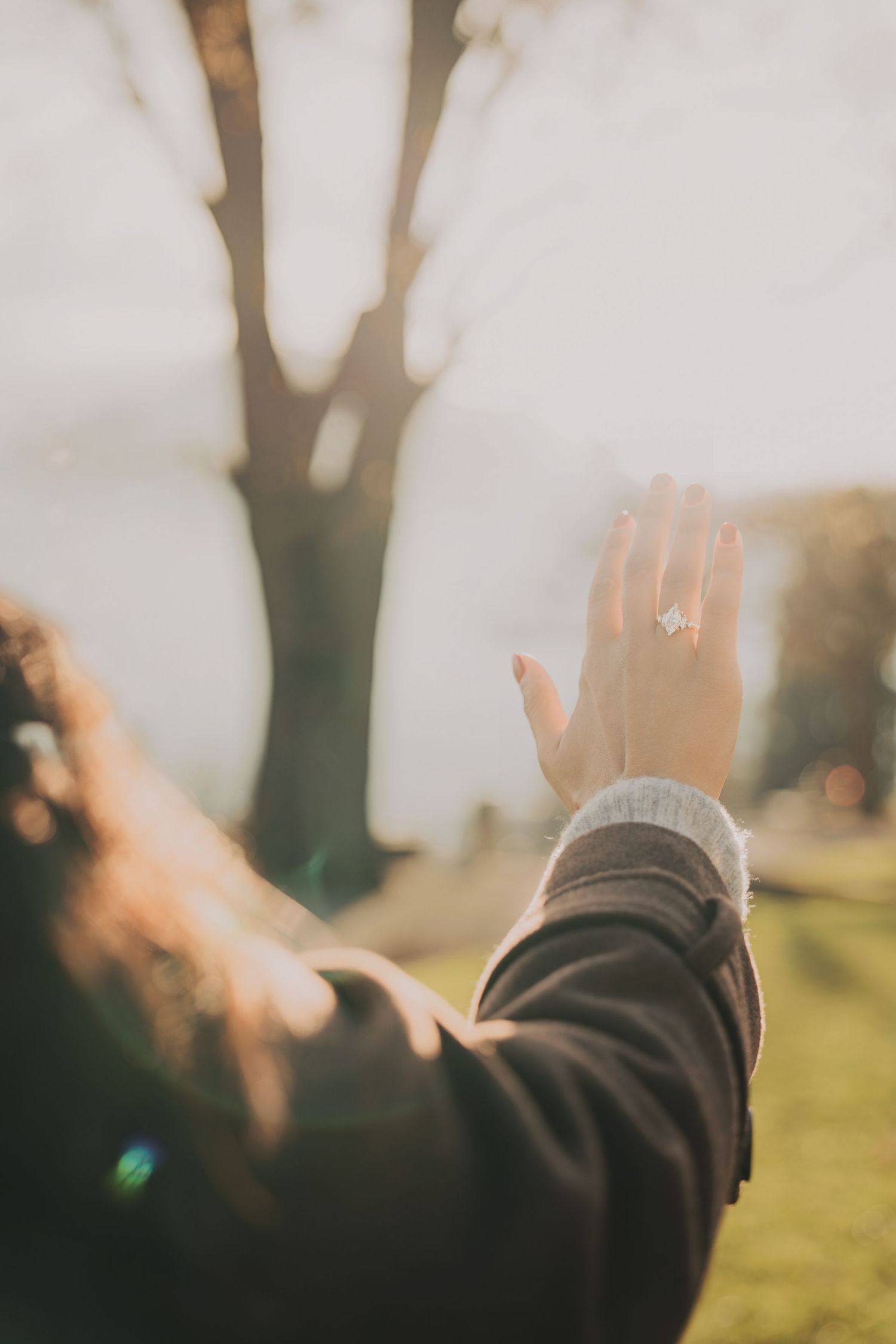 Woman raising hand to display diamond engagement ring with sunlit tree and park in background.
