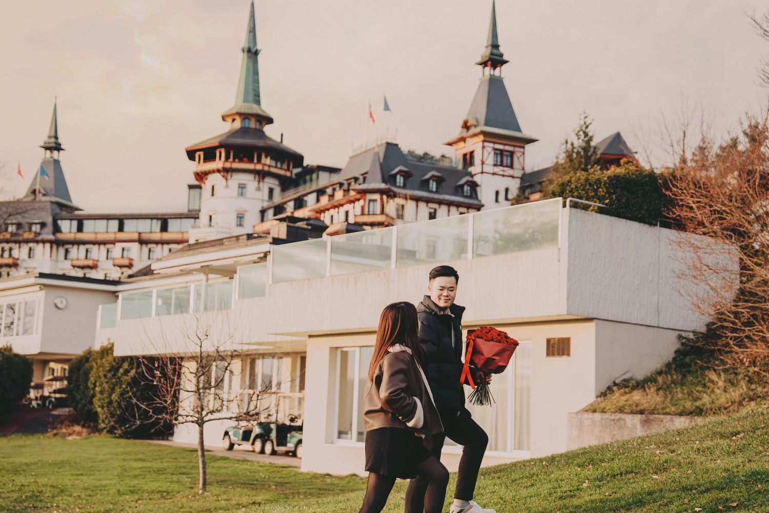 Couple playfully walking on green lawn with grand historic castle hotel behind them.