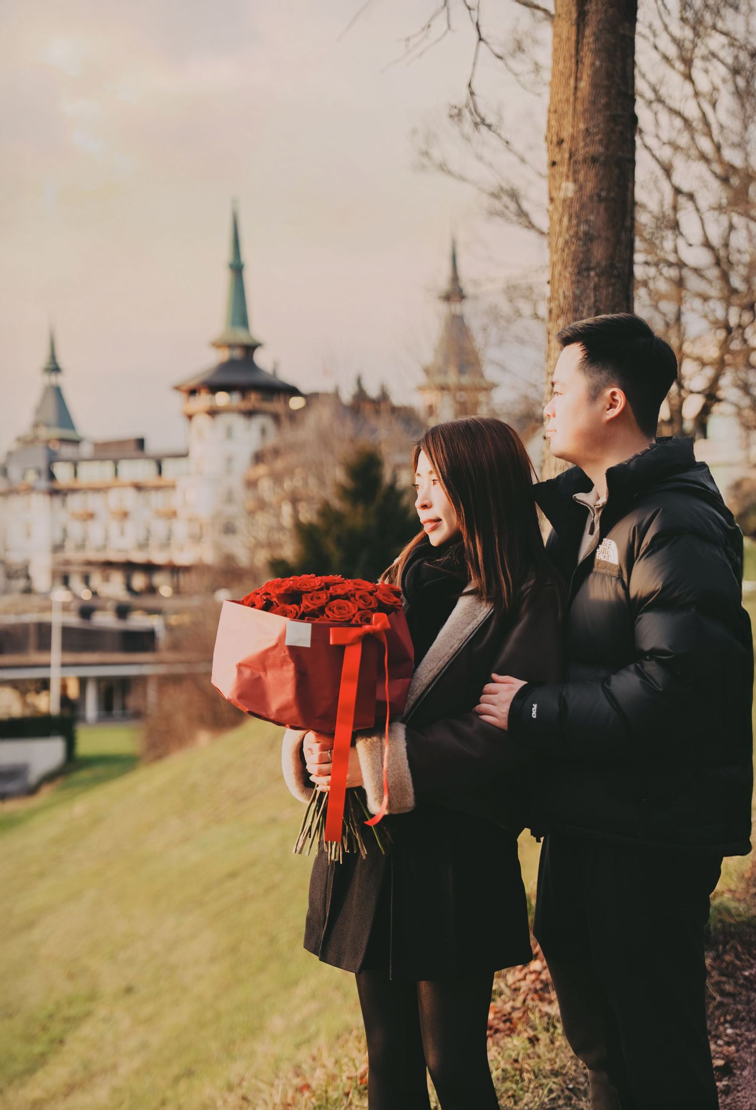 Couple sharing romantic moment near tree holding large red rose bouquet before castle.