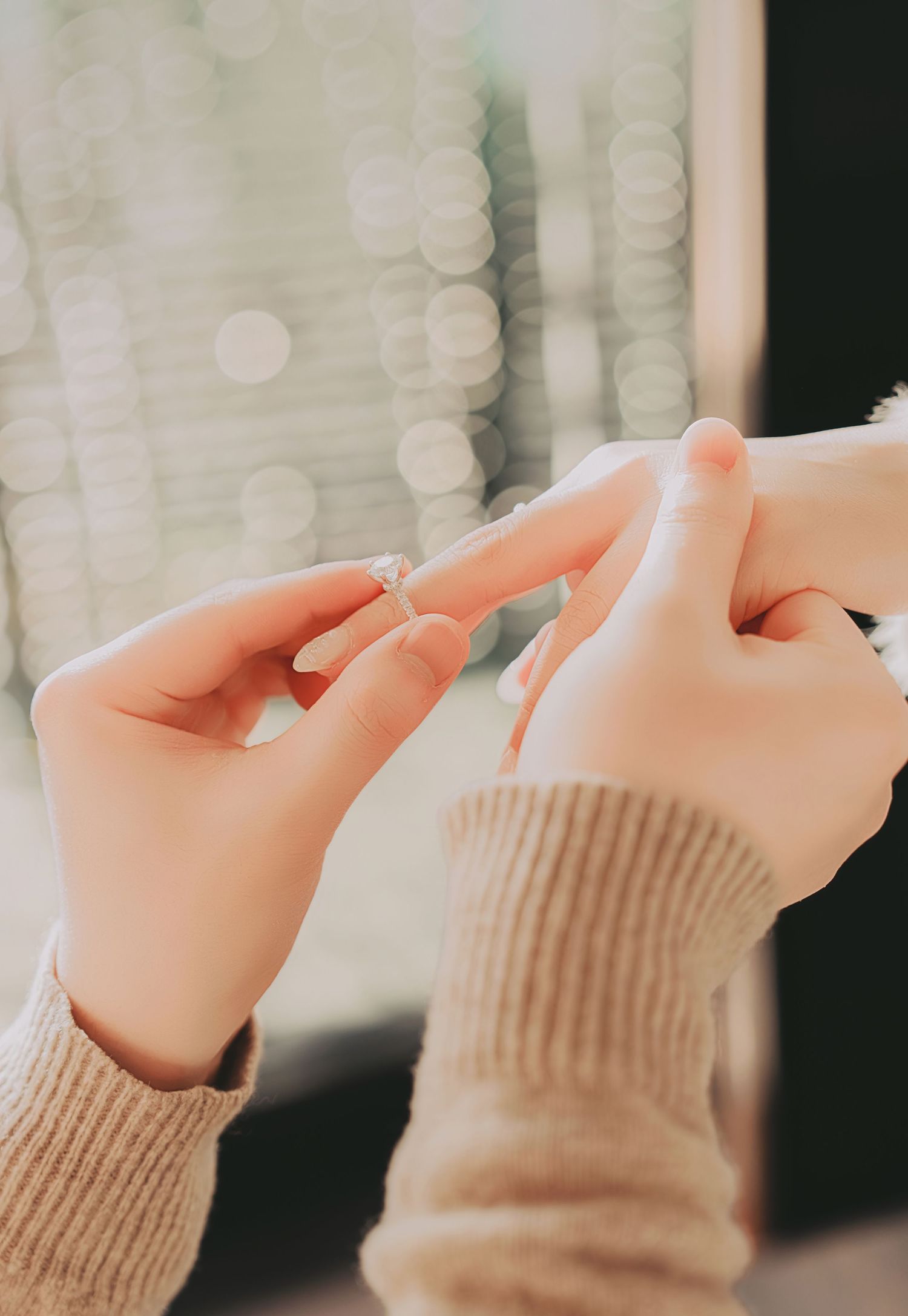 Tender moment of an engagement ring being placed on a woman's finger with soft bokeh lights in background.