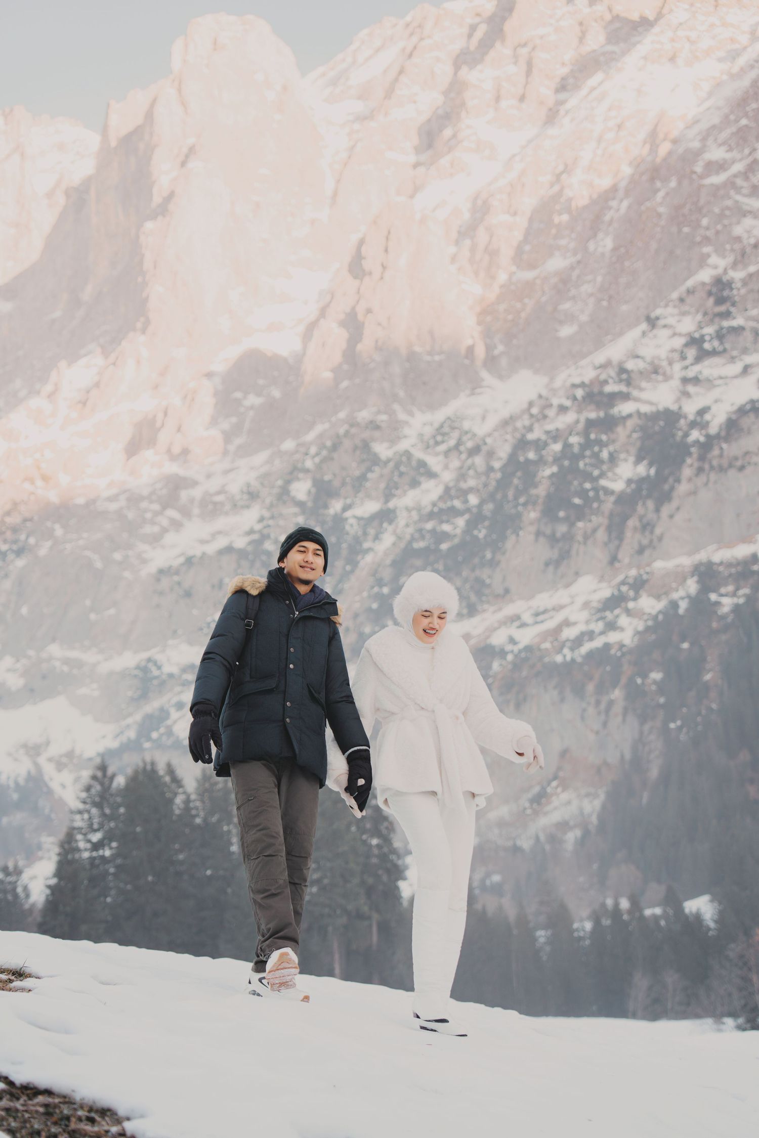 Couple walking through snow with towering rocky mountains and pine trees in the background.