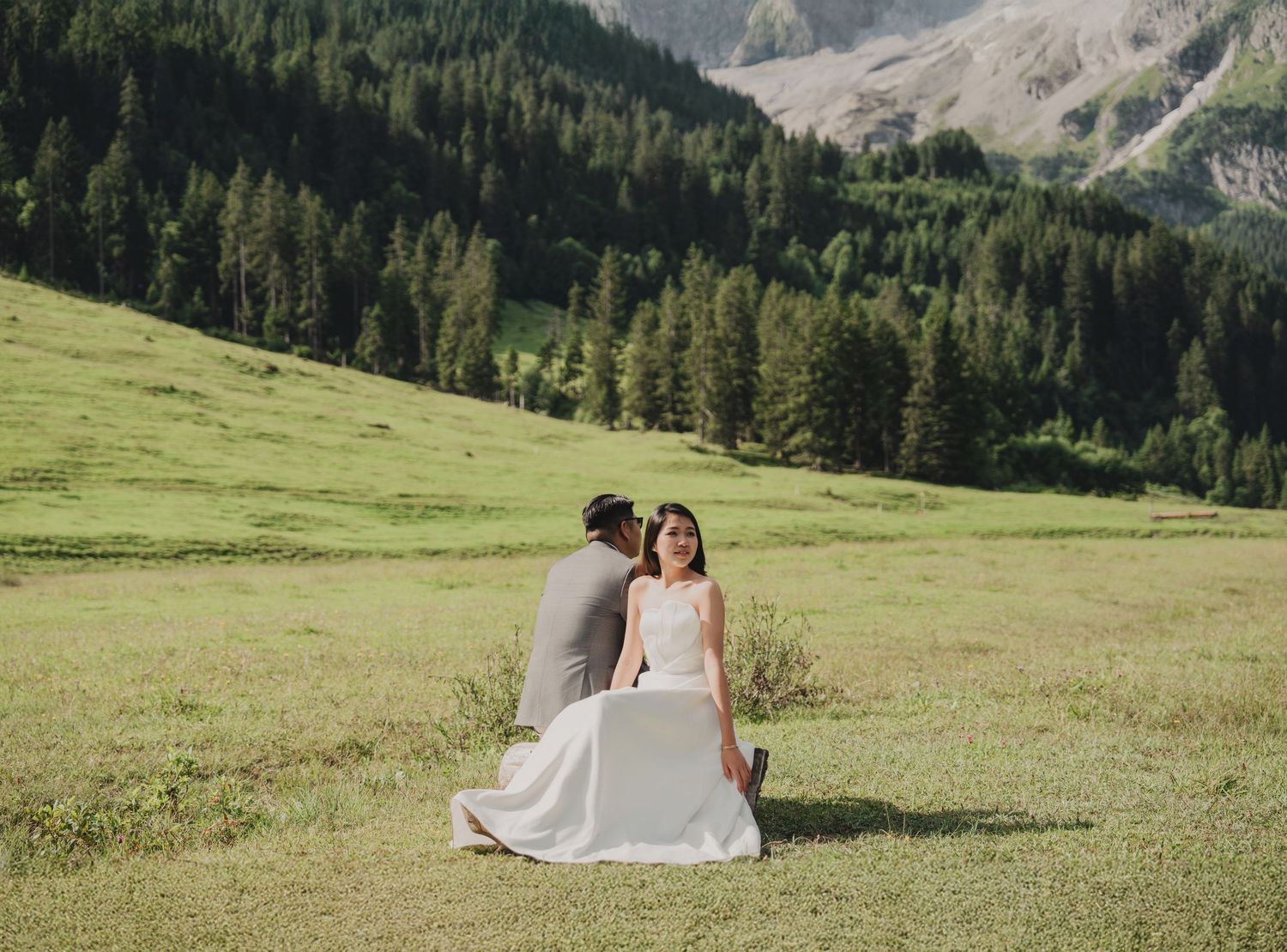 Groom whispering to seated bride in white dress on green meadow with mountain forest backdrop.