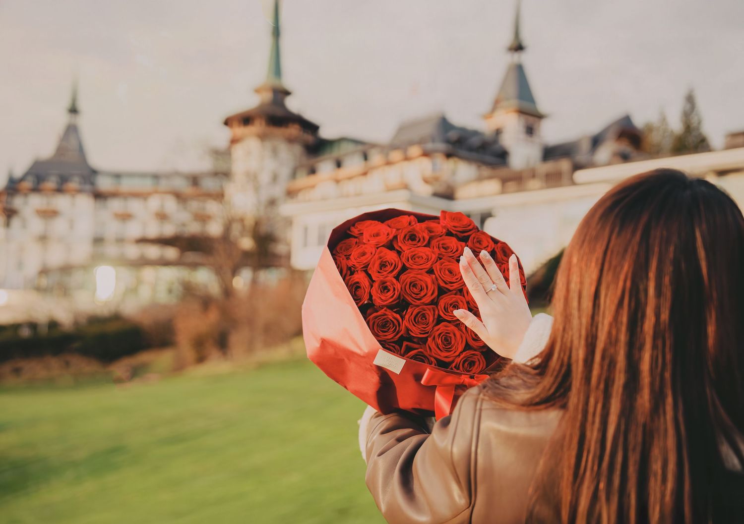 Woman holding heart-shaped box of red roses in front of grand European castle building.
