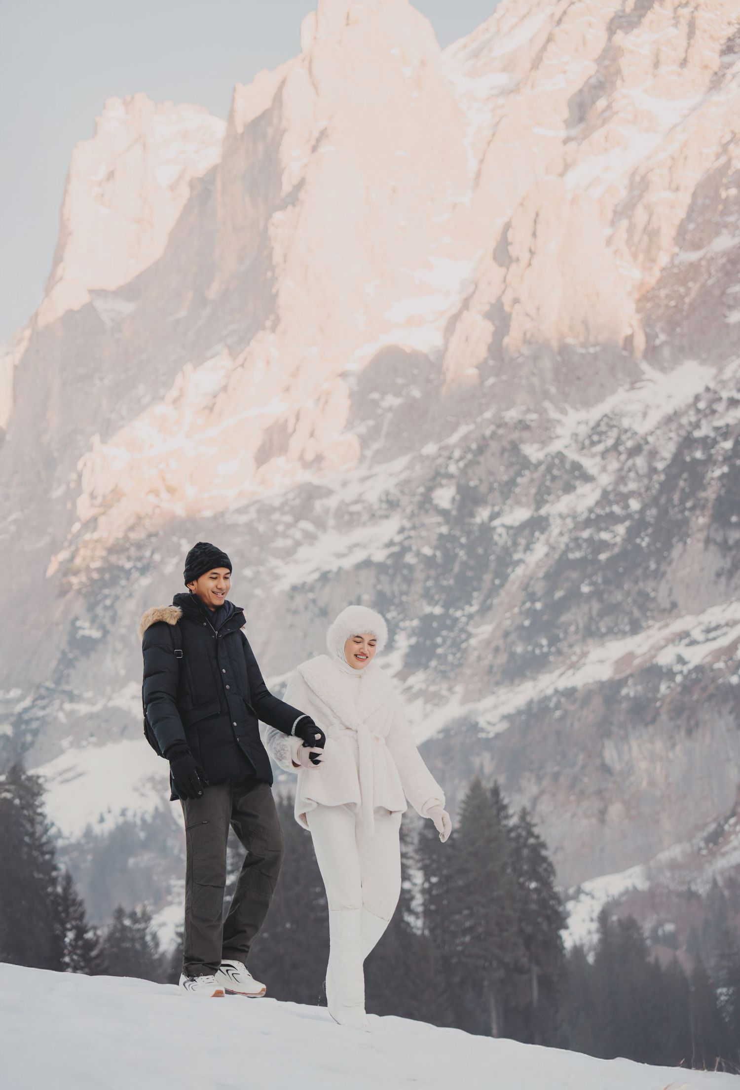 Couple walking in snow with dramatic mountain backdrop, woman in white hijab and man in dark jacket holding hands.