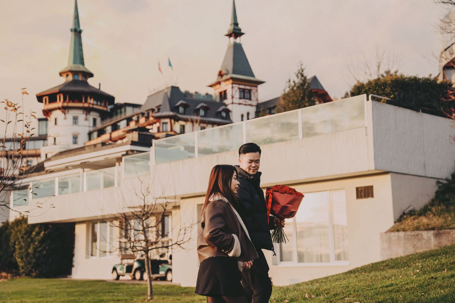 Couple walking together near modern white building with historic castle hotel behind them.