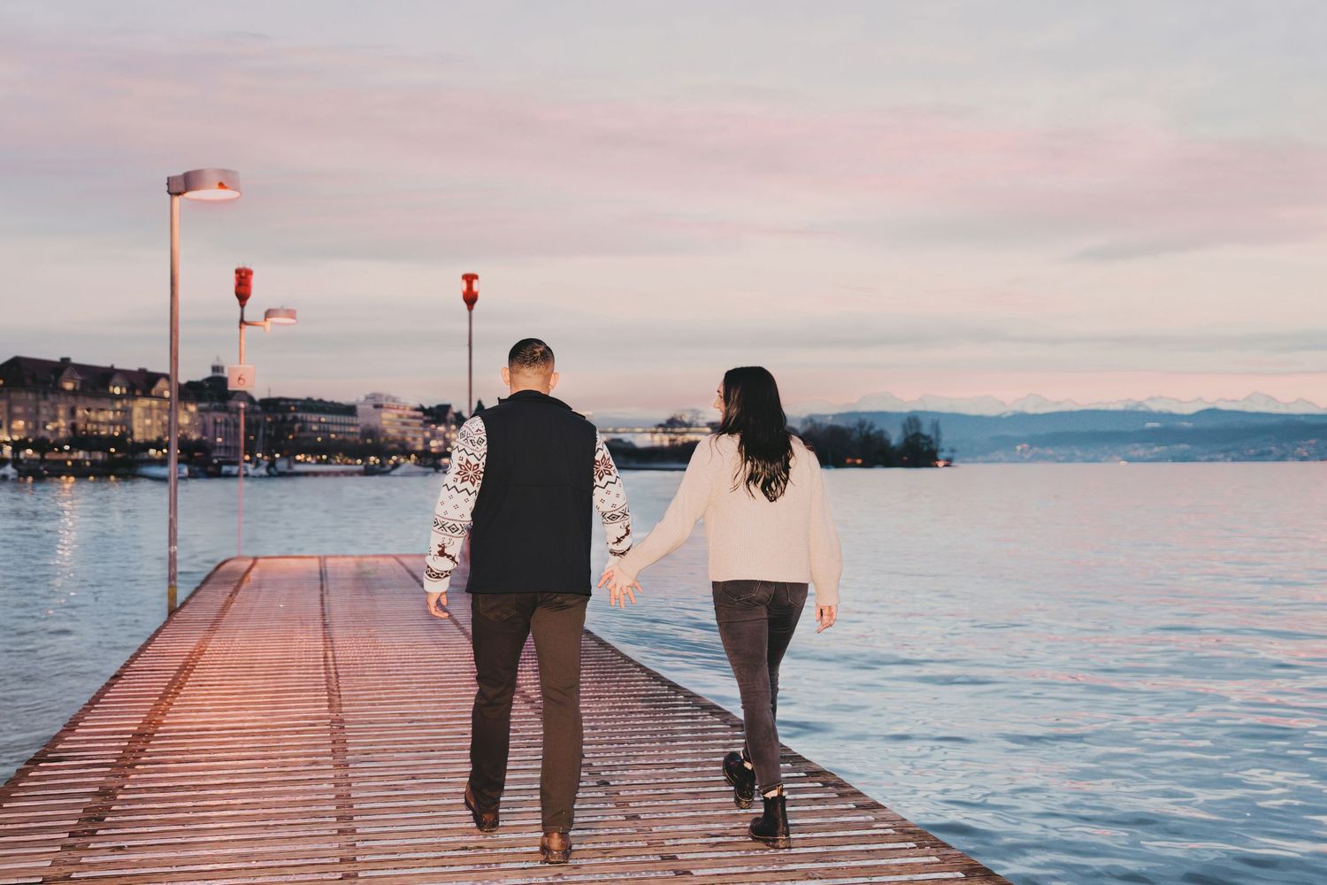 Couple walking hand in hand along a wooden pier at sunset over calm water.