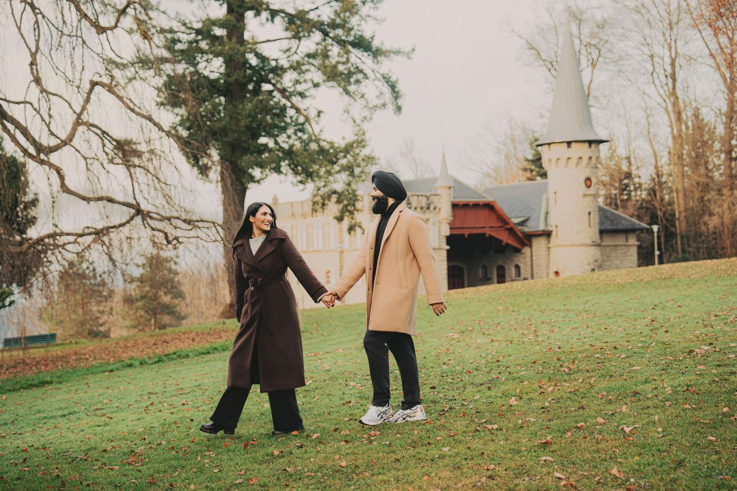 Couple holding hands and smiling at each other on a green lawn with a fairytale castle backdrop.
