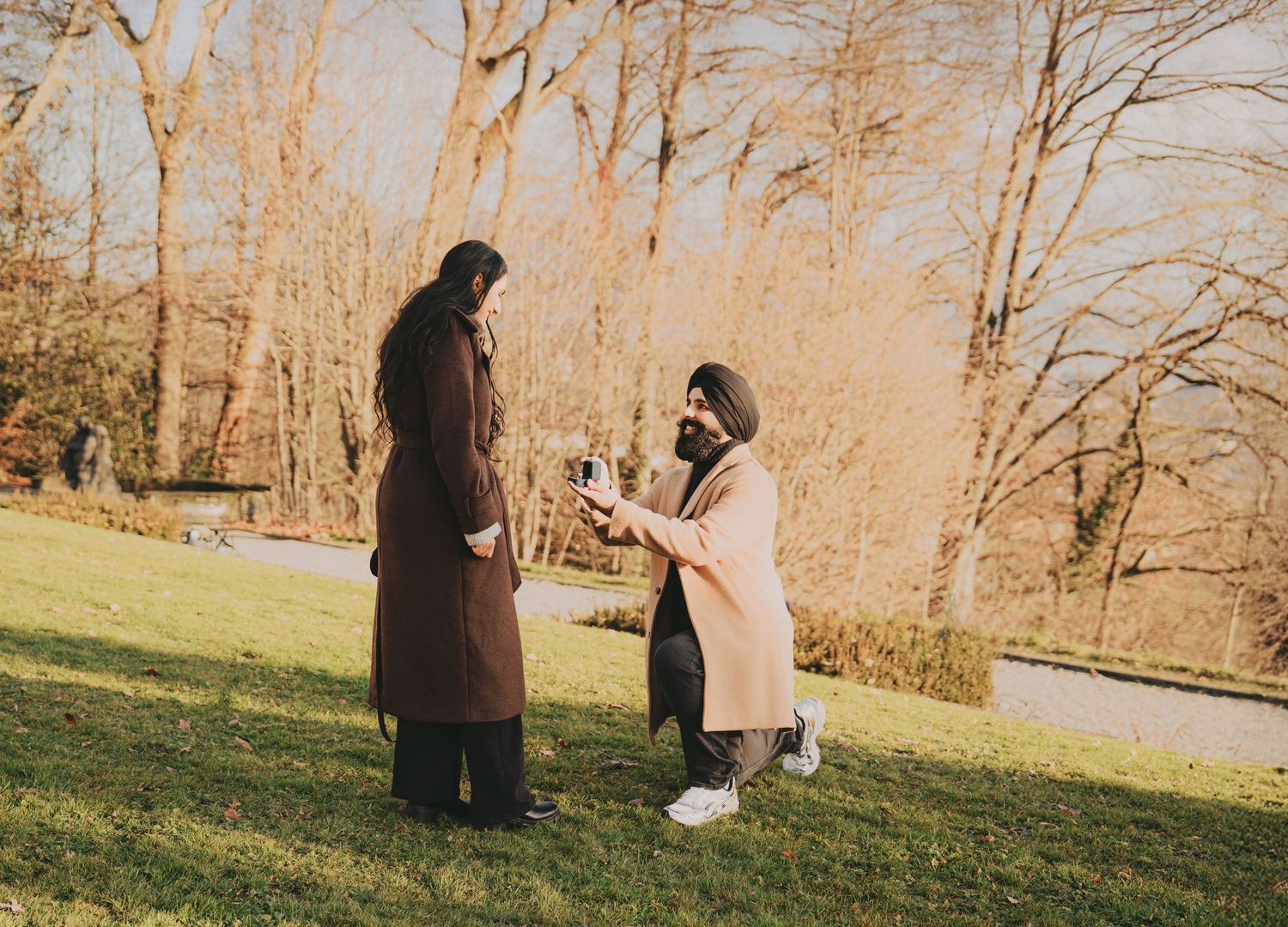 Sikh man kneeling on one knee proposing to woman in brown coat in autumn park setting.
