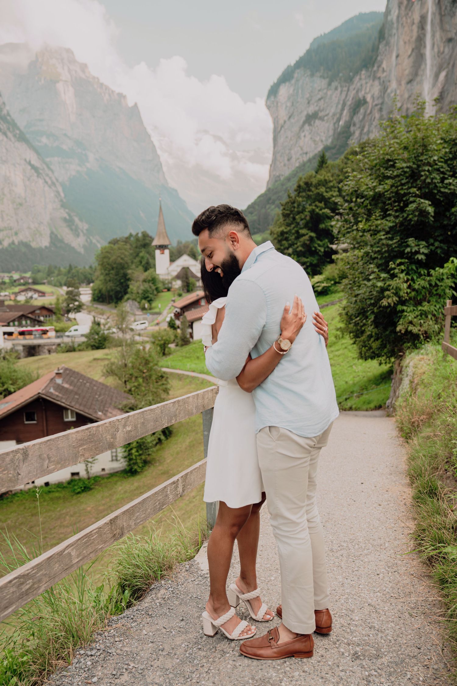 Couple embraces on a scenic mountain path in a Swiss alpine village after a proposal.