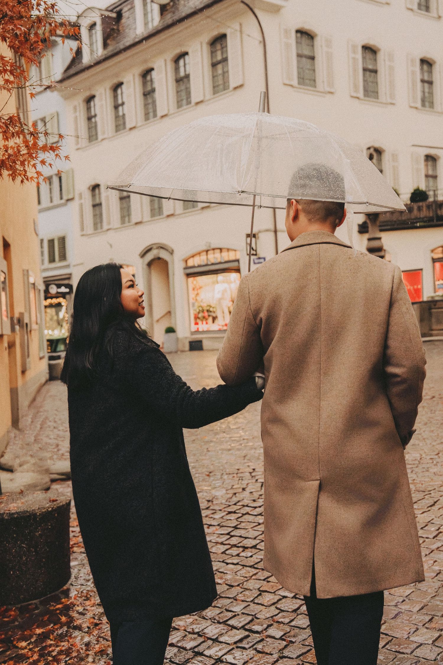 Couple walking cobblestone streets of Zurich in autumn, sharing a clear umbrella in the rain.