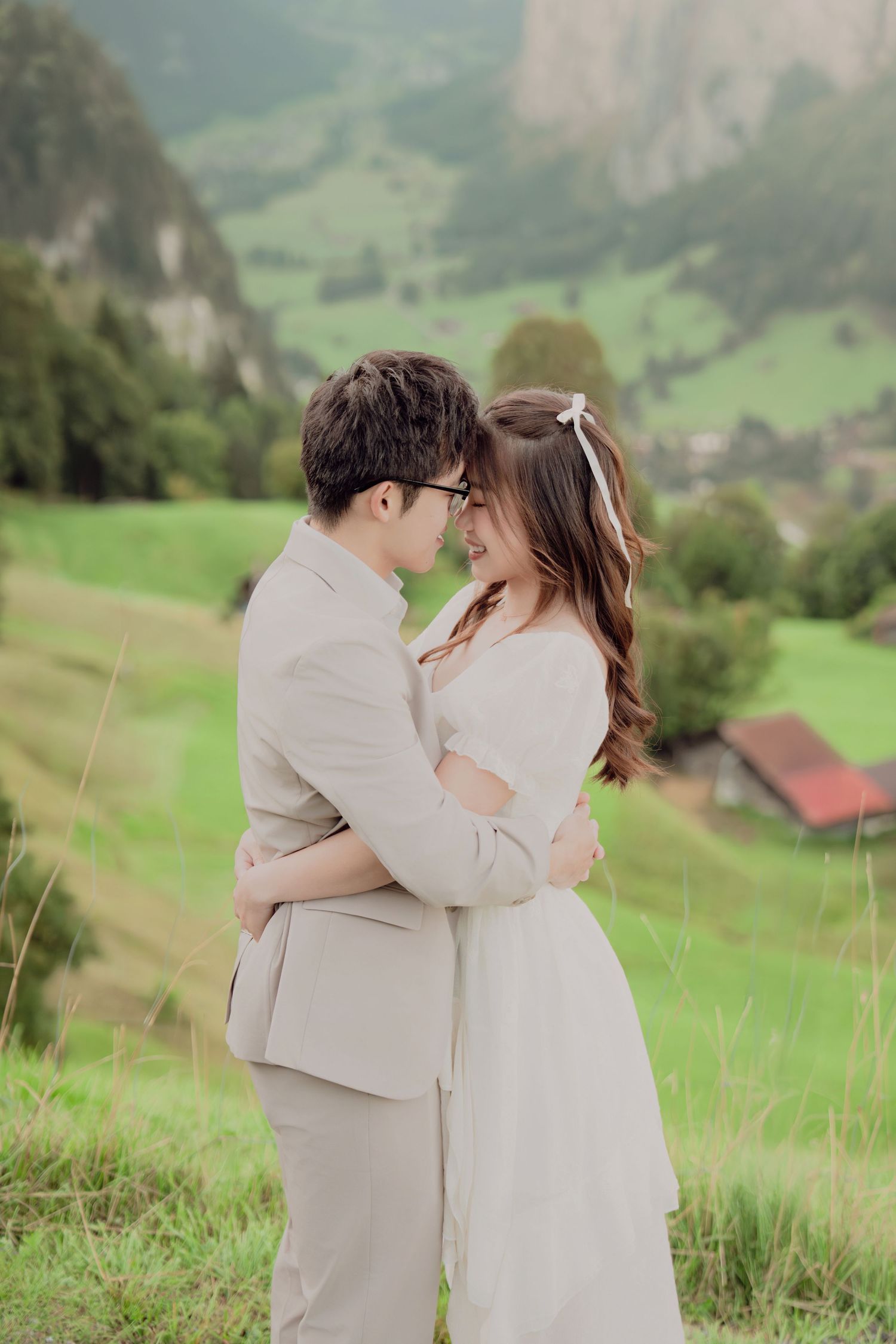 Romantic couple holding each other close smiling on lush green Swiss alpine meadow with mountain village.