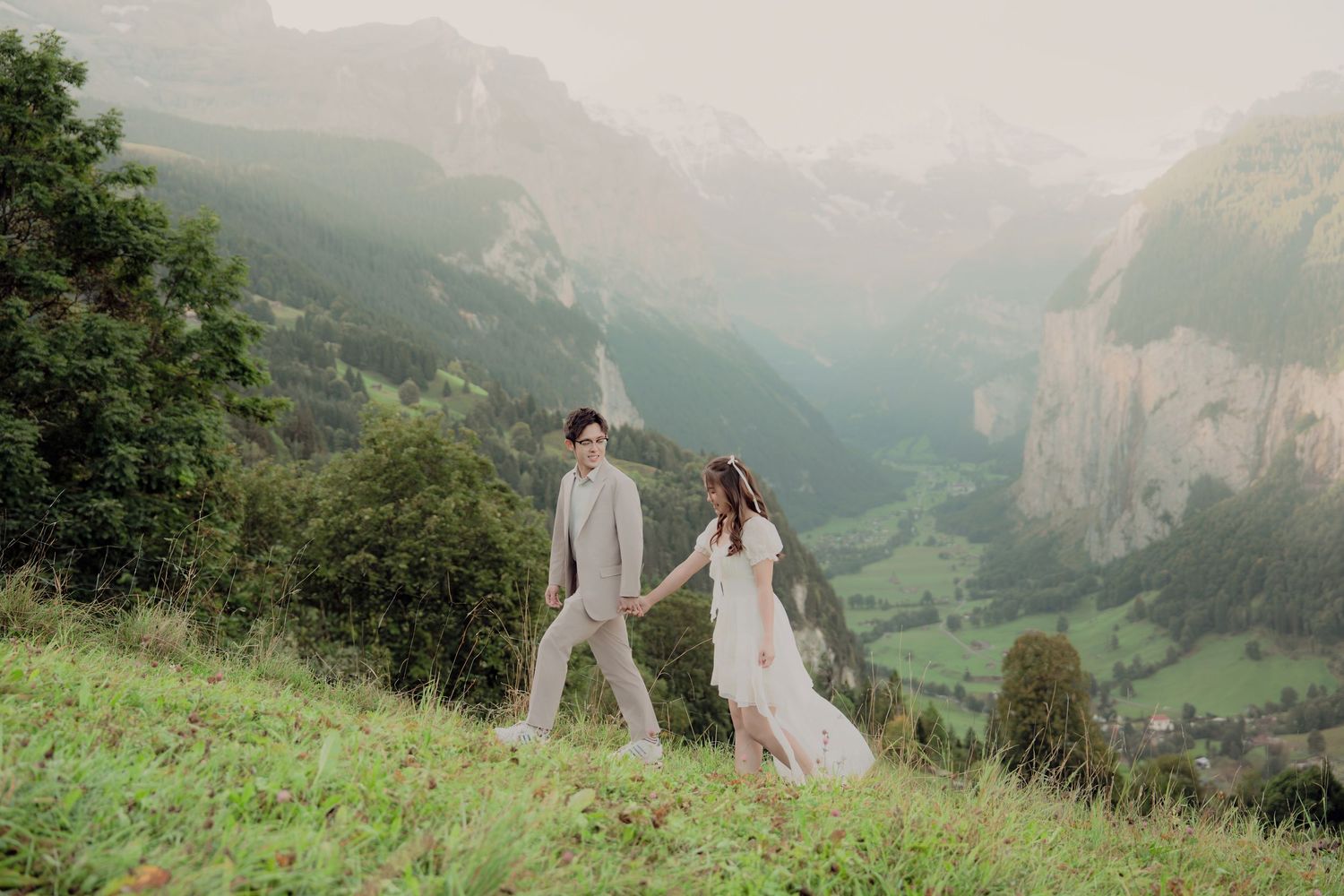Couple holding hands walking on a green hillside with a stunning Swiss Alpine valley and misty mountains behind them.