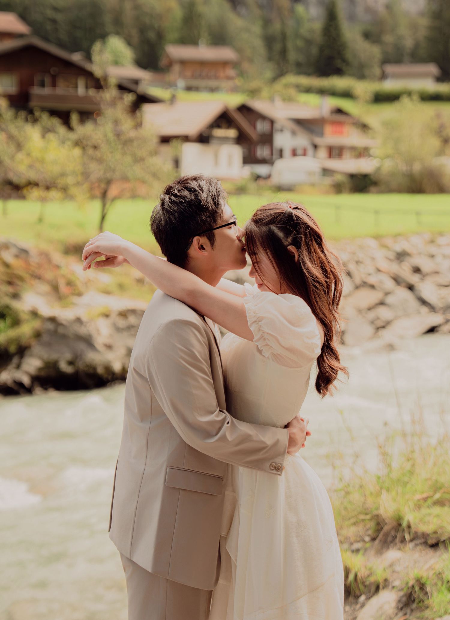 Couple sharing a romantic kiss by a river with Swiss chalet village in the background.