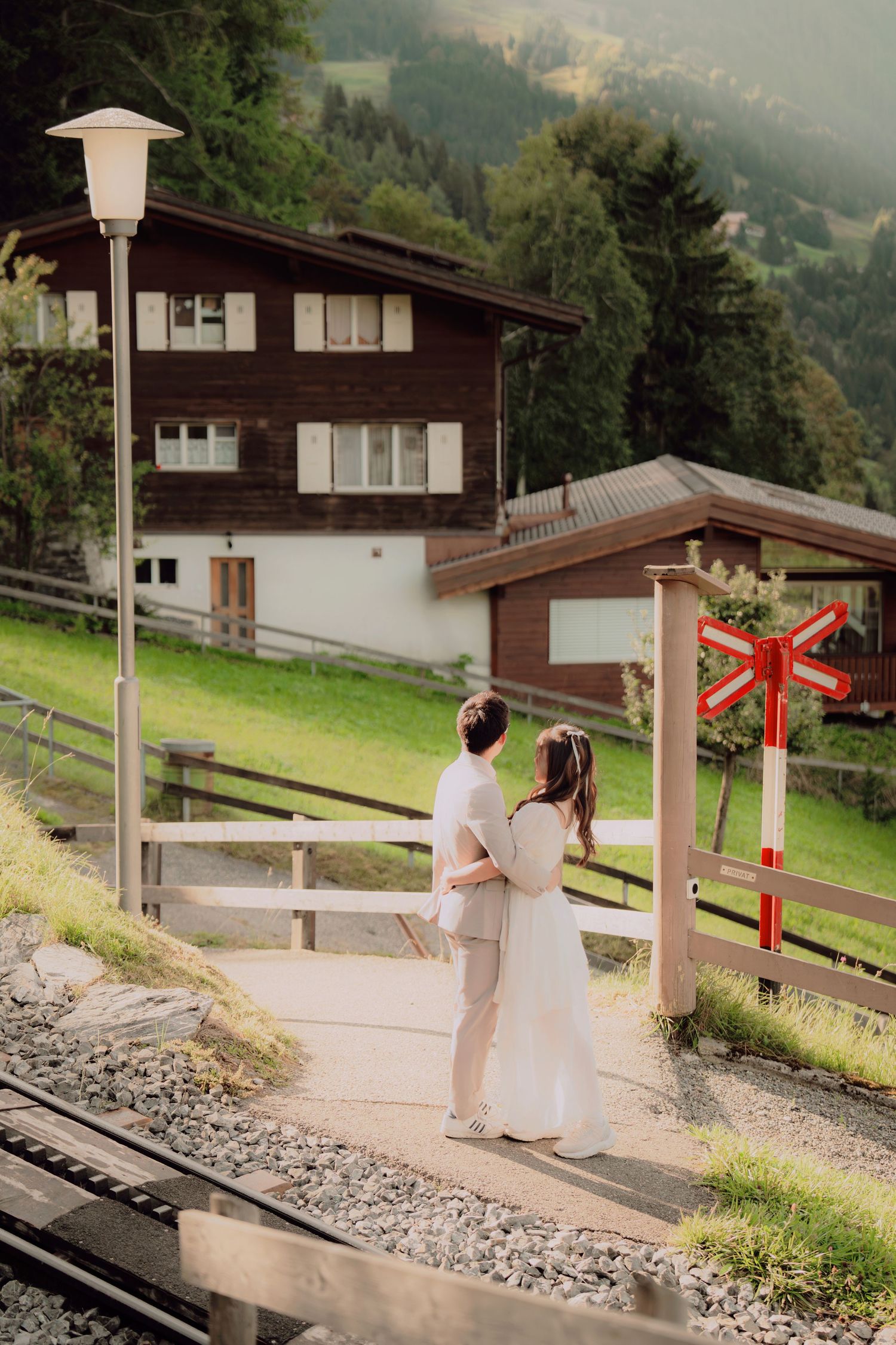 Wedding couple stands together at a mountain train station crossing with a red signal sign and alpine chalet backdrop.