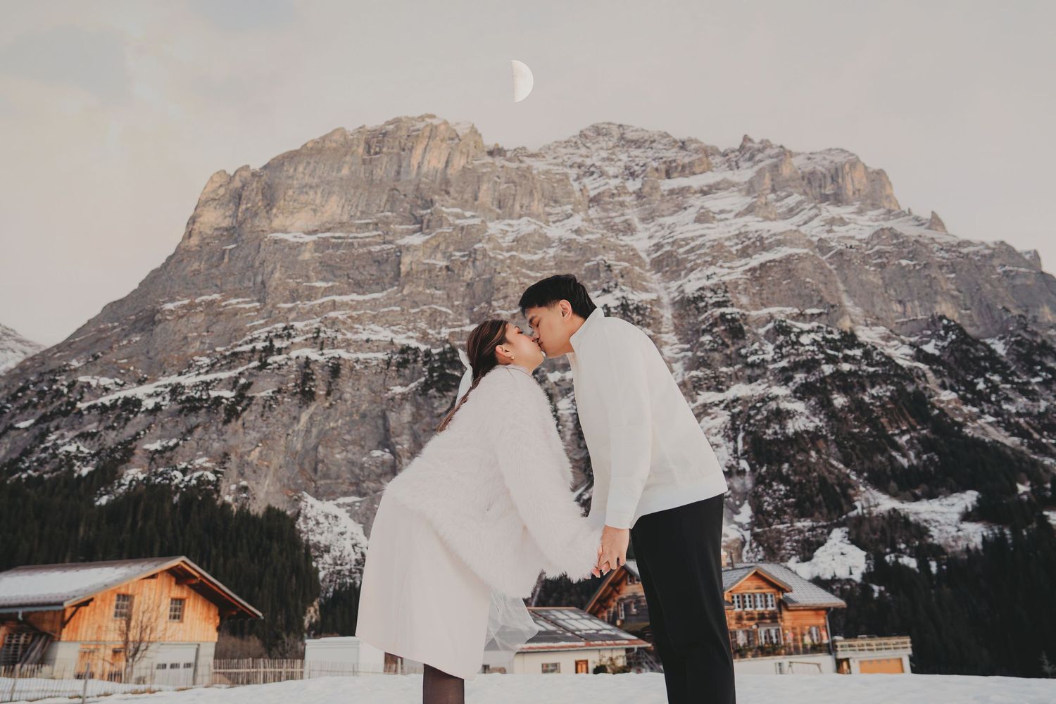 Couple kissing passionately in snow with dramatic snowy mountain and crescent moon overhead.