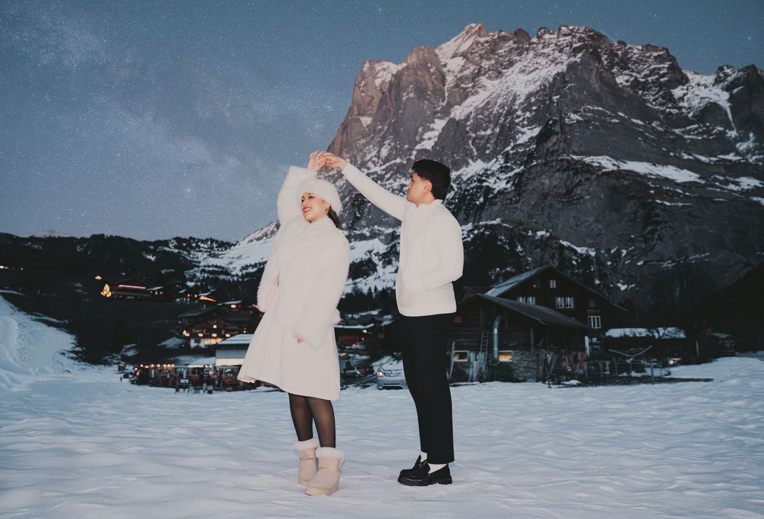 Couple dancing at night in snowy mountain village under starry sky in white outfits.