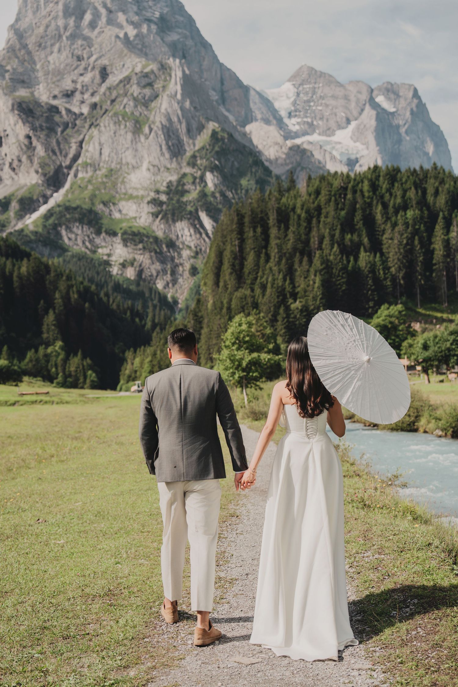 Wedding couple walking hand in hand on mountain path, bride holding white parasol umbrella.