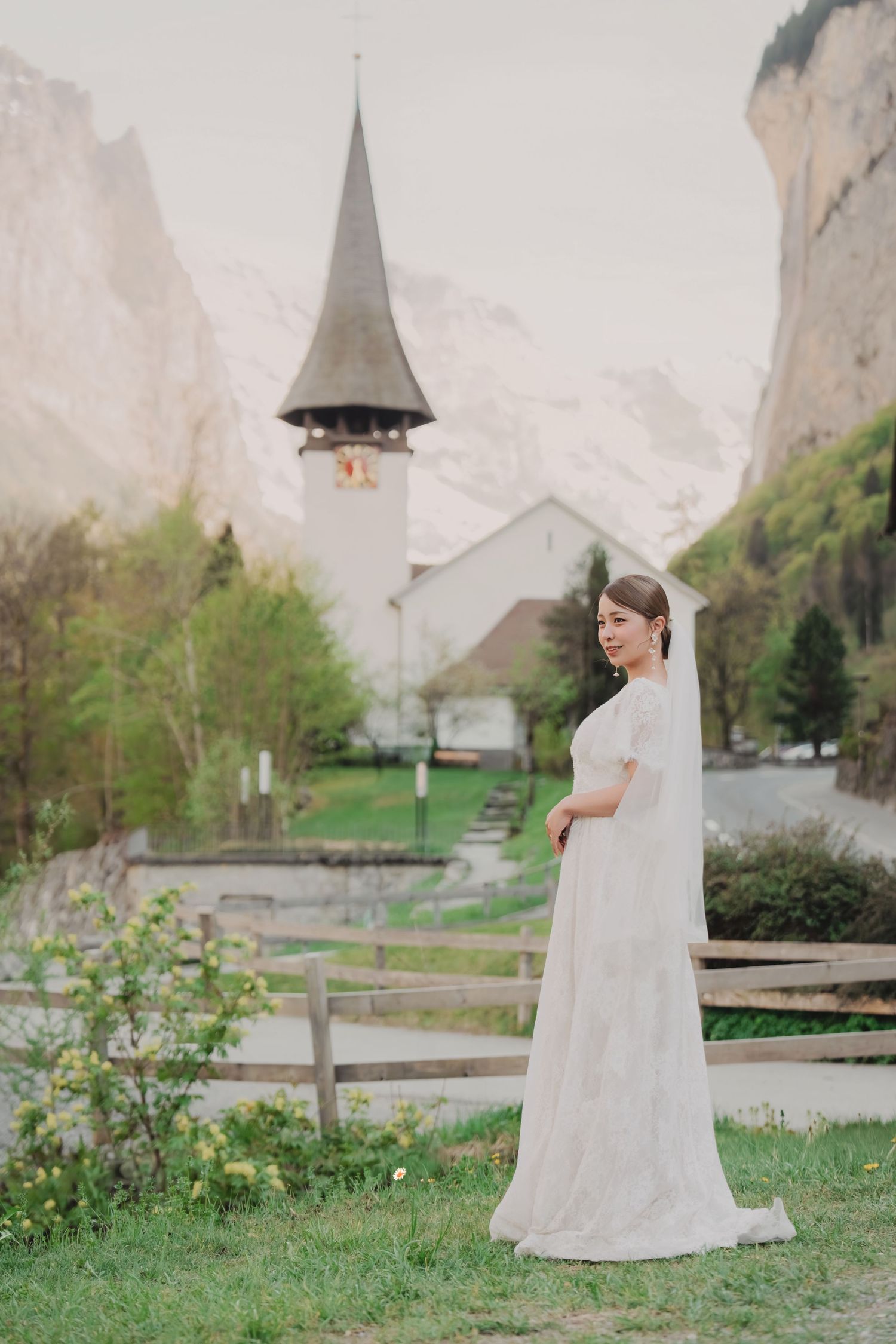 Elegant bride in lace wedding gown stands before a Swiss alpine church in Lauterbrunnen.