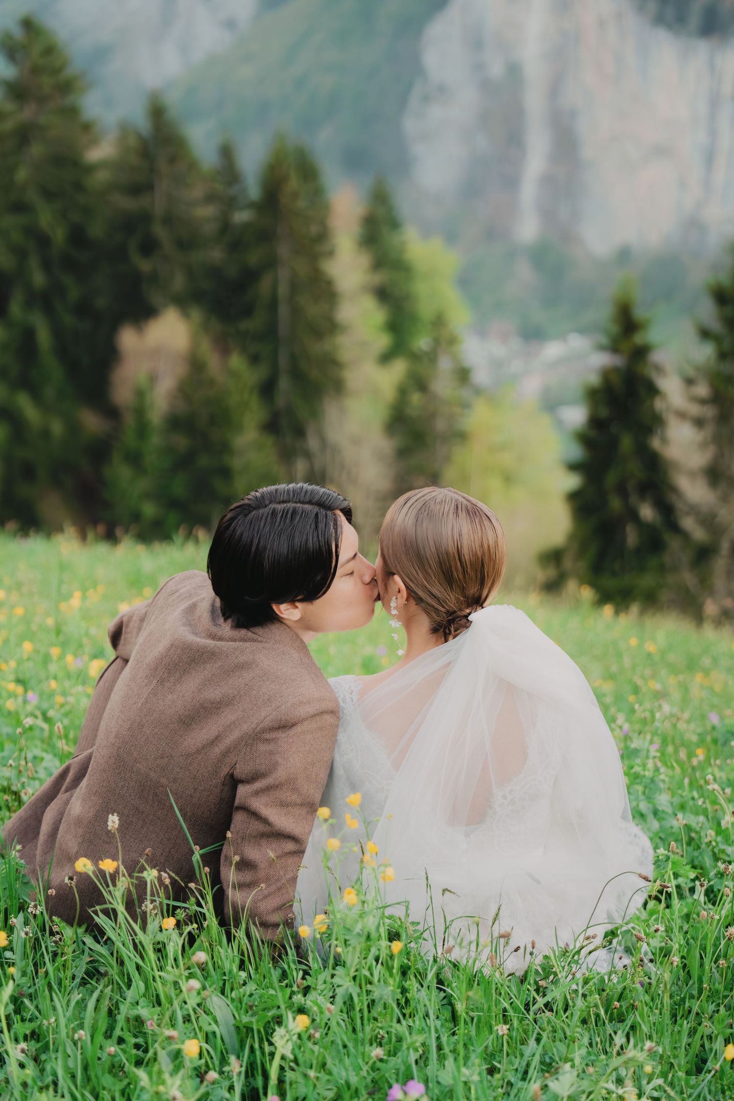Wedding couple sharing a kiss while sitting in alpine wildflower meadow with mountain backdrop.