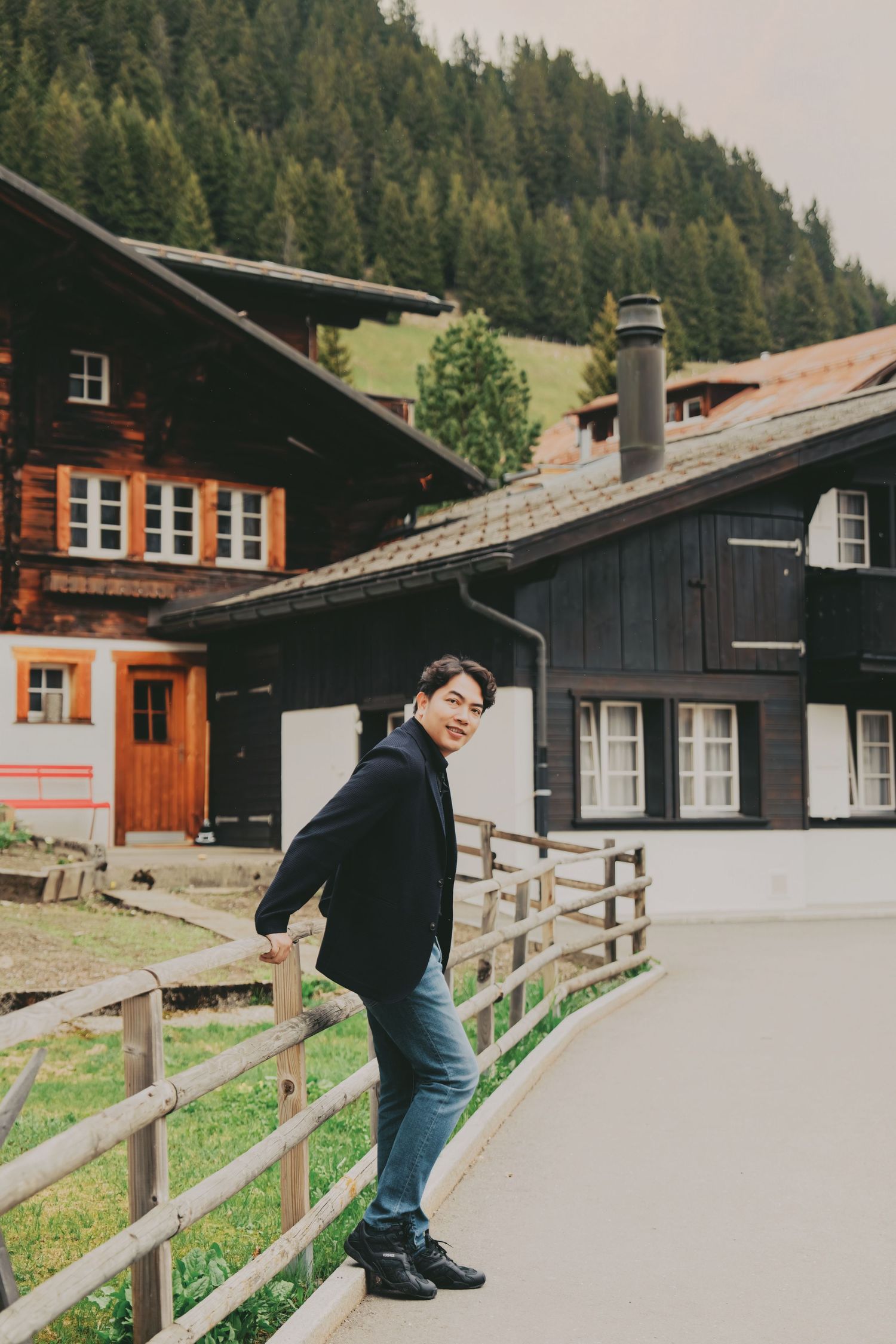 Smiling Asian man leaning on wooden fence beside traditional Swiss alpine chalets with dense forest behind.