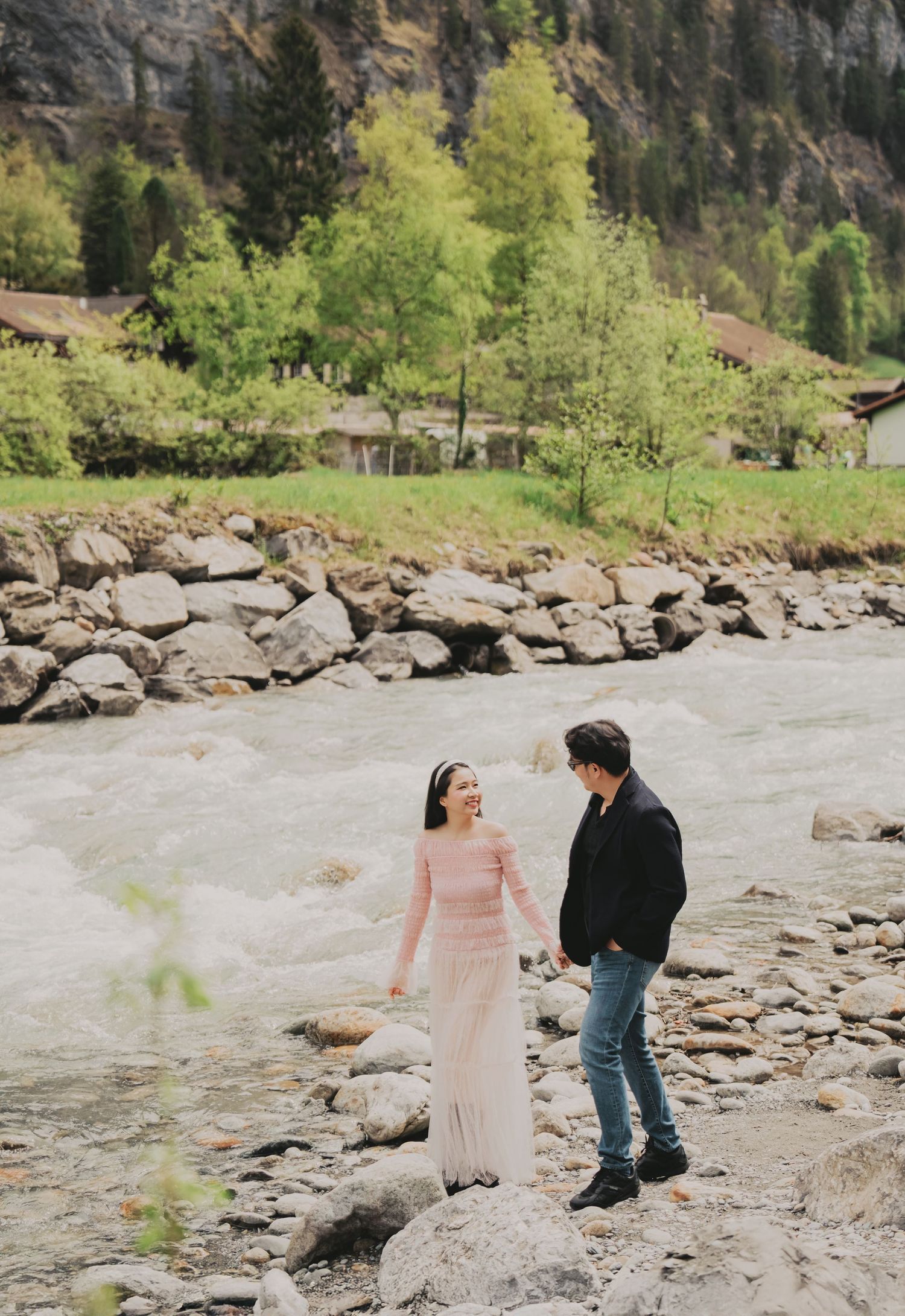Happy couple holding hands and laughing together on rocky shore beside a swift mountain river.