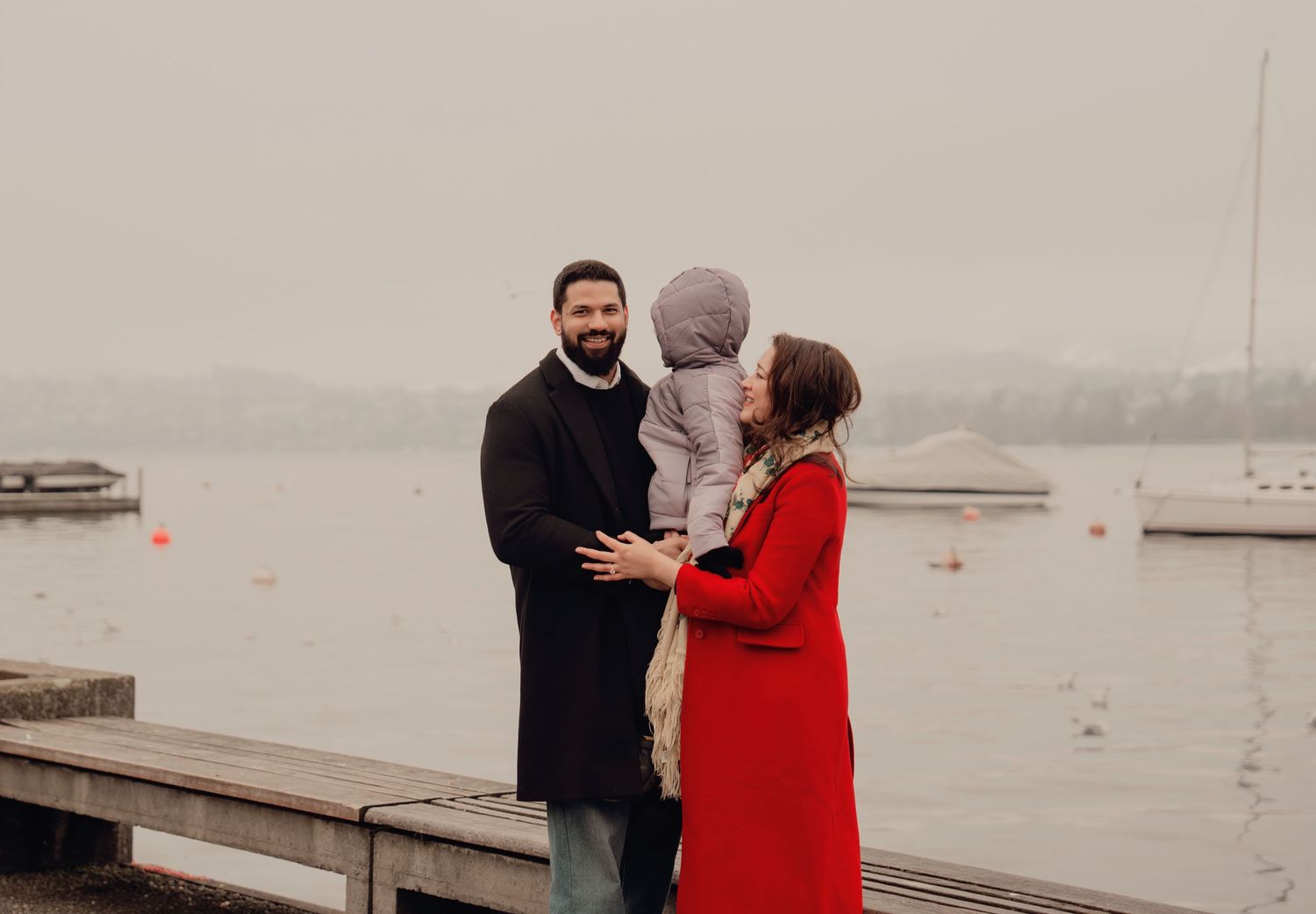 Family of three standing by a misty lakeside, father holding child in gray hat, mother in red coat.